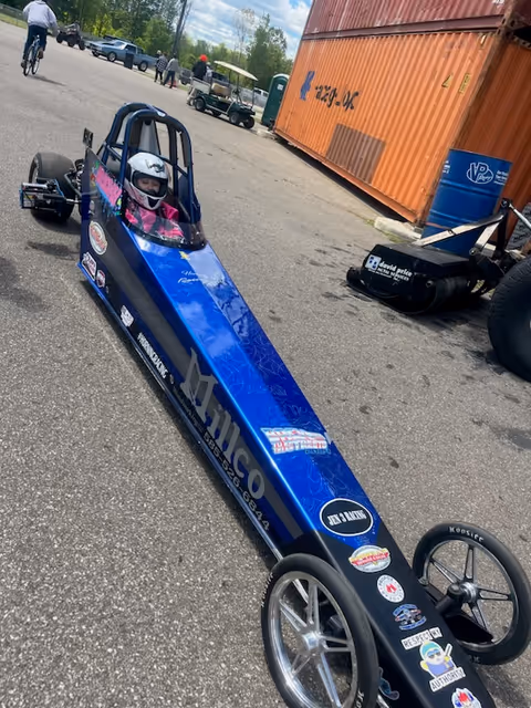 A blue dragster with a helmeted driver parked outdoors.