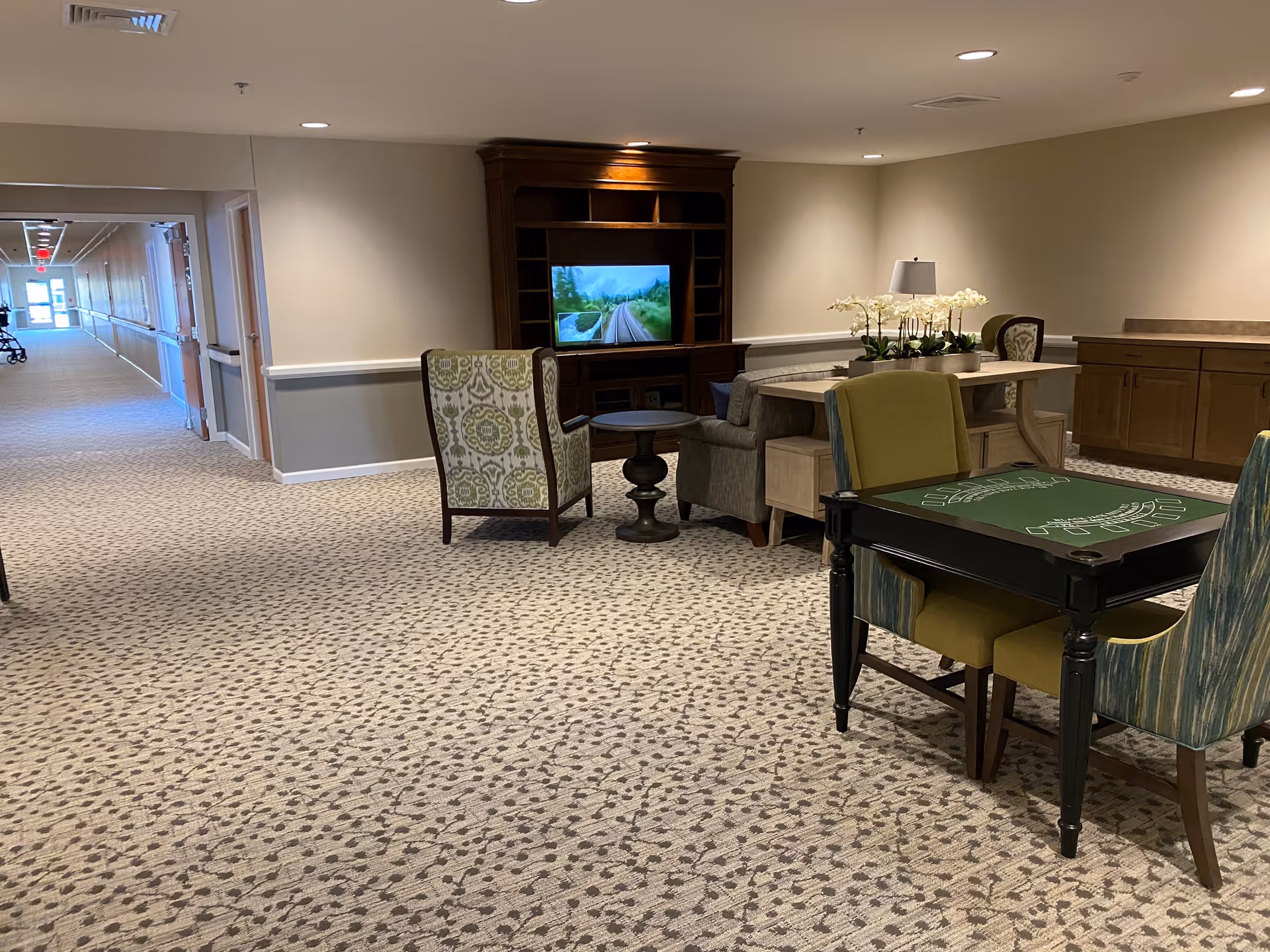A cozy common area in a senior living facility with patterned carpet, a wooden entertainment center with a TV, several upholstered chairs, a small round table, a rectangular table with a lamp and flowers, and a card table with four chairs. A hallway extends to the left with handrails along the walls.