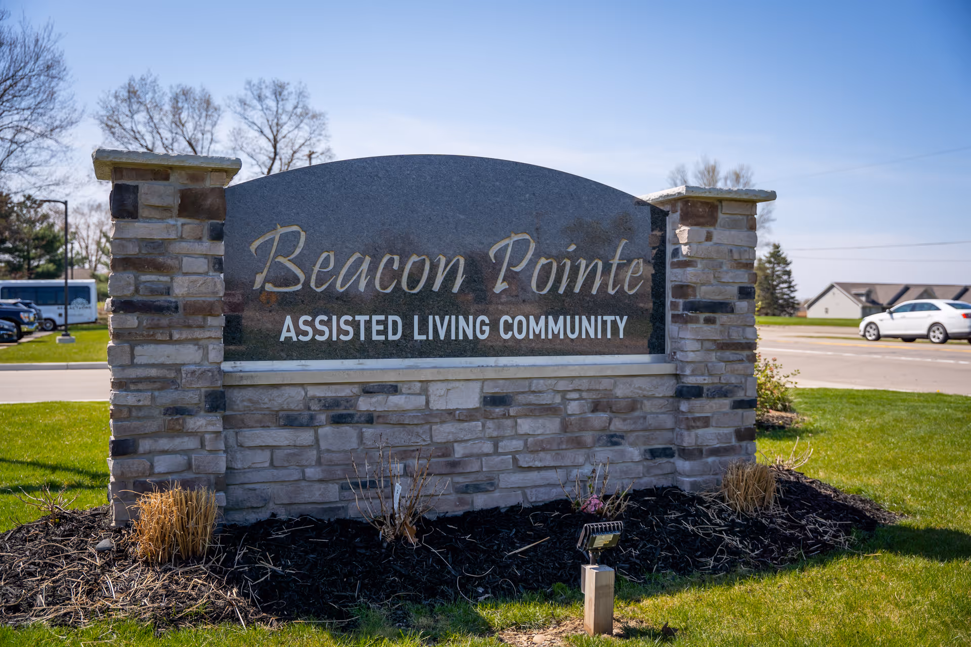 Stone sign for Beacon Pointe Assisted Living Community set on a grassy area near a road with cars and trees in the background under a clear blue sky.