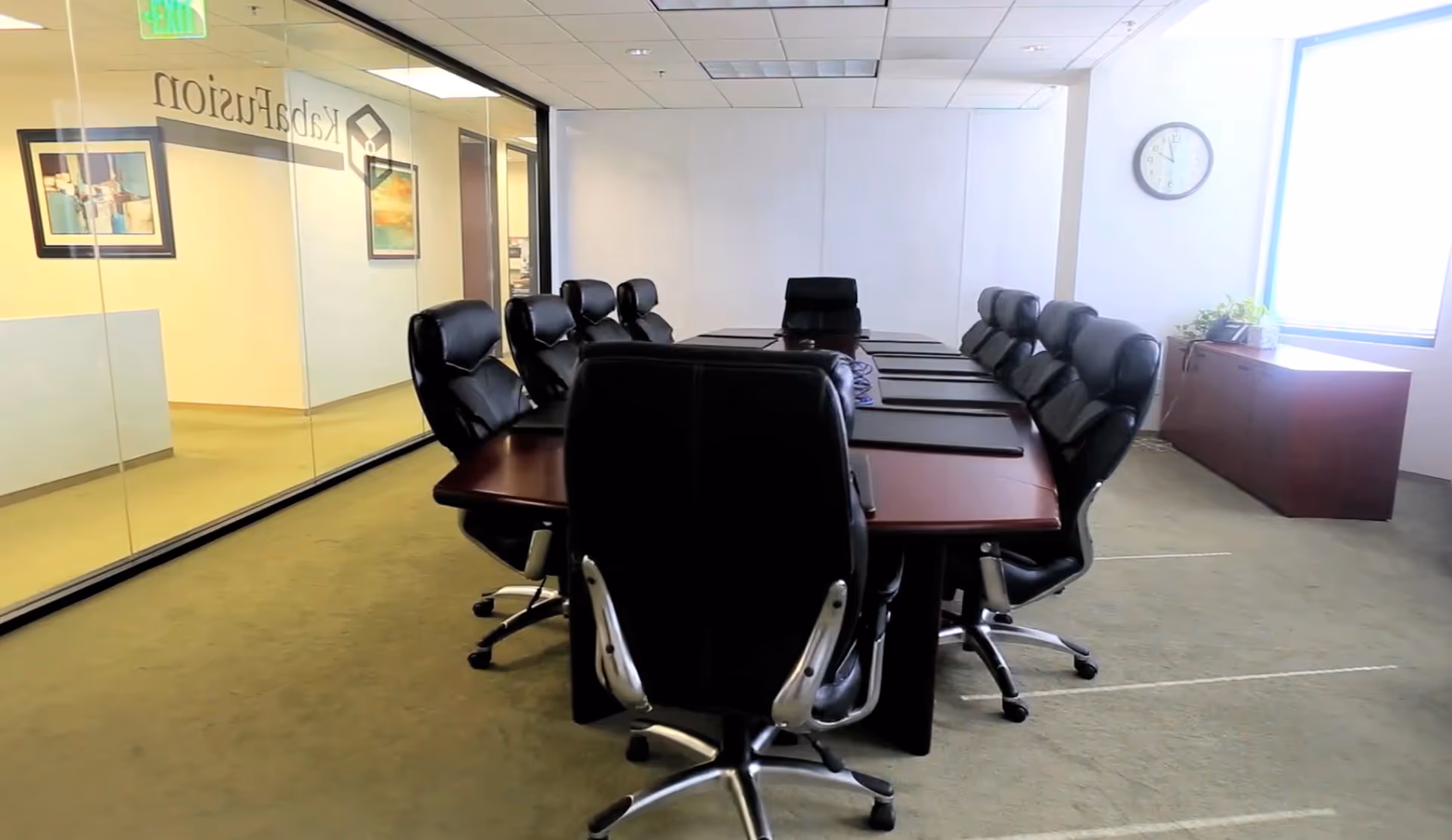 A conference room with a long wooden table, black executive chairs, and glass walls.