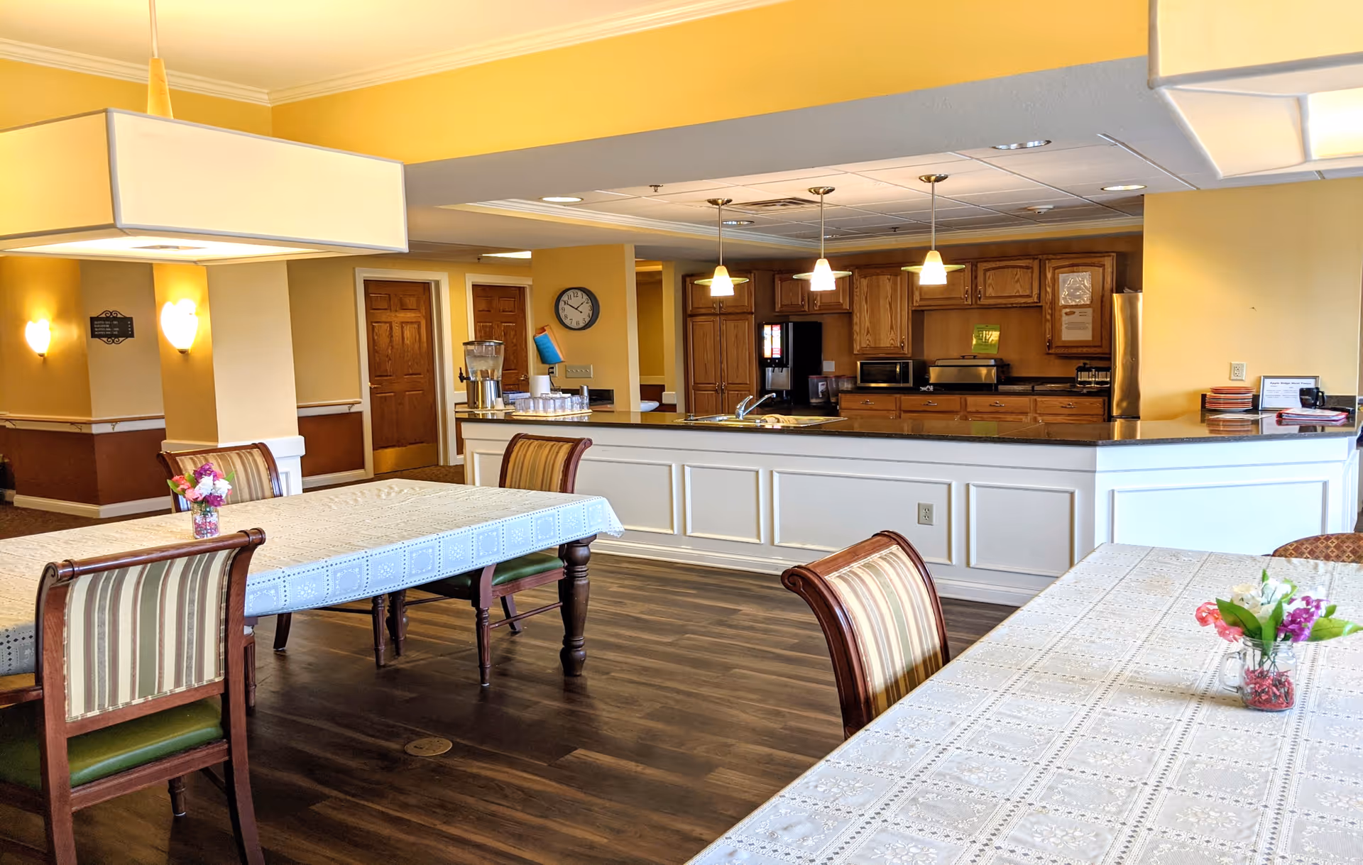 A dining area in a senior living facility featuring tables covered with white patterned tablecloths and small flower arrangements. The room has wooden chairs with striped upholstery, wood flooring, and a kitchen area in the background with wooden cabinets, a refrigerator, microwave, and a counter with a sink. Pendant lights hang from the ceiling above the counter.