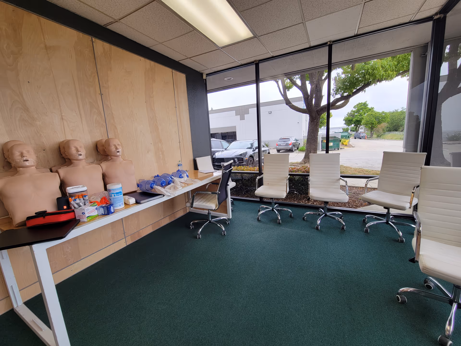 A CPR training room with lifelike mannequins, medical supplies, and white chairs by a large window.