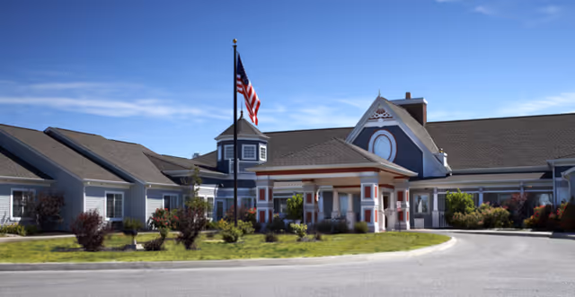 Front exterior of a single-story senior living facility with a covered entrance, American flag, and landscaped lawn under a blue sky.