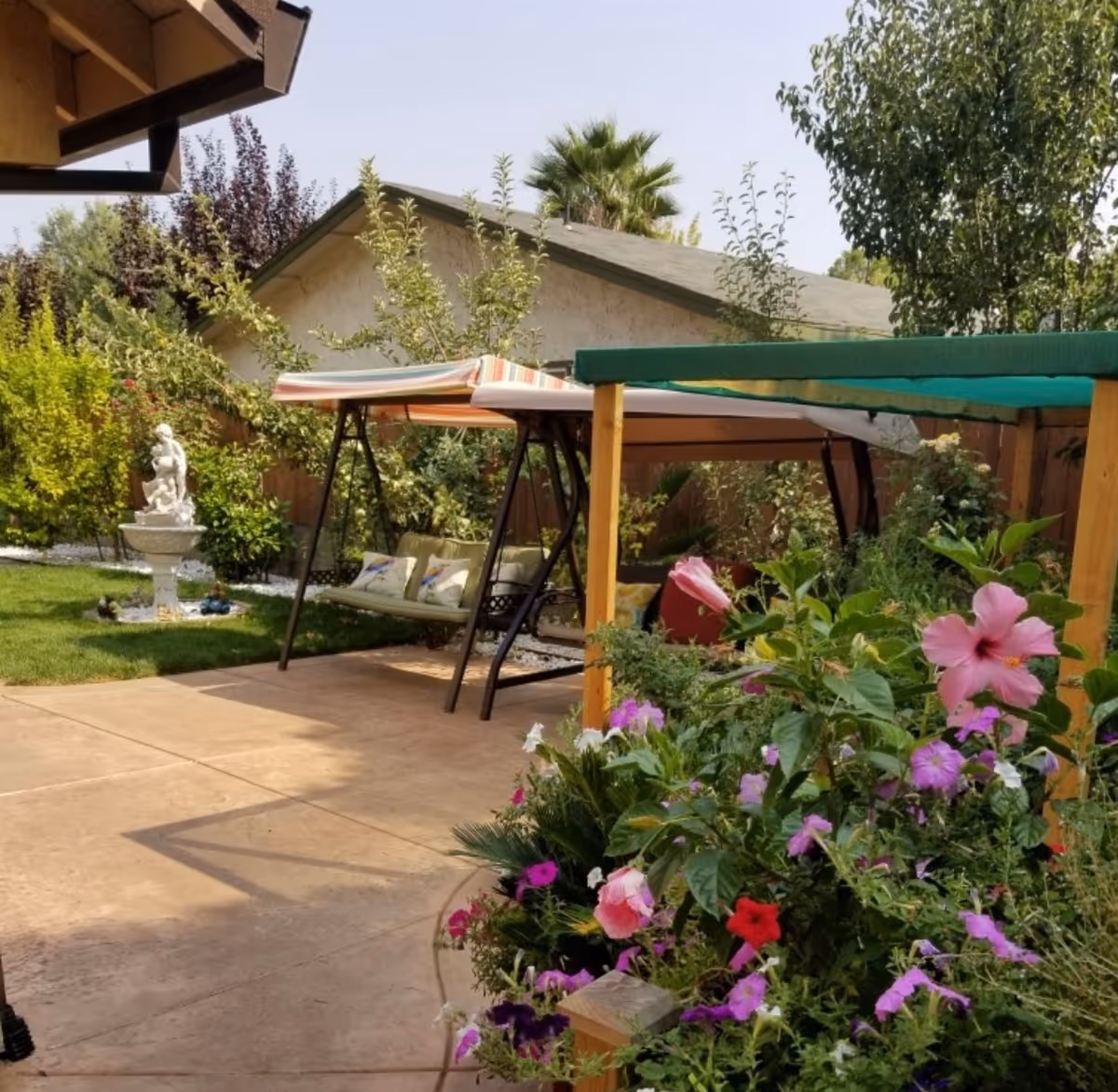 Sunny outdoor patio with a garden, swing bench, shade canopies, and blooming flowers.