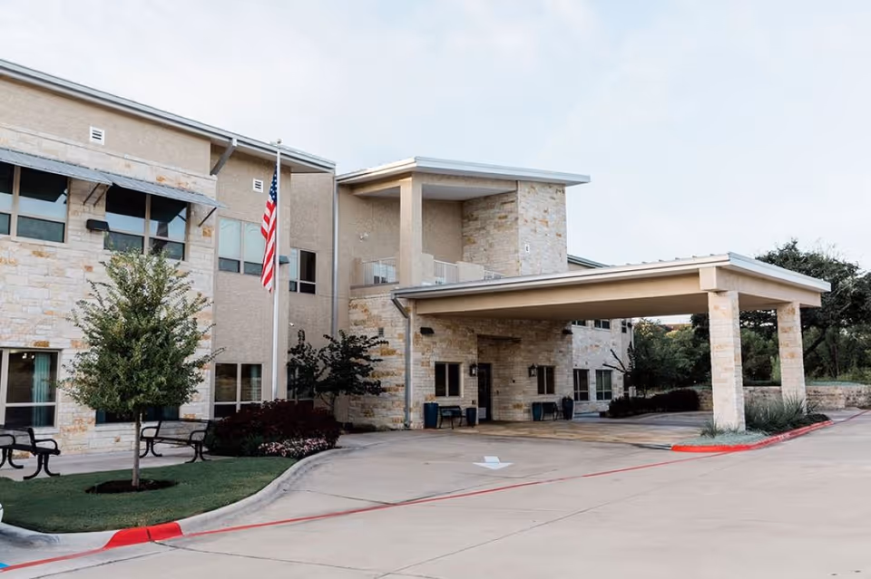 Exterior view of a senior living facility building with stone and beige walls, a covered entrance driveway, an American flag on a flagpole, benches, small trees, and landscaped greenery.