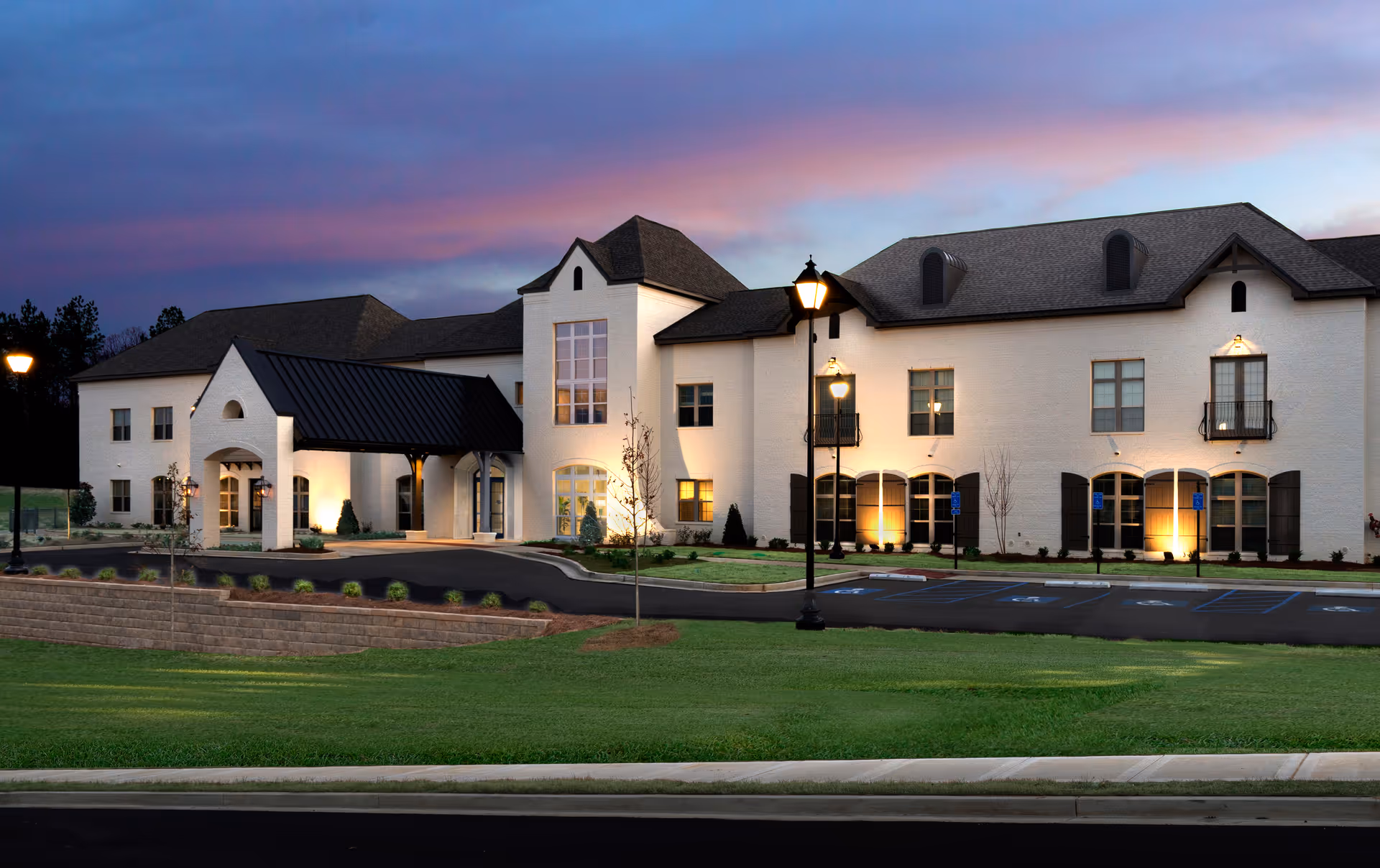 Front exterior of a light colored senior living facility with lamps and parking.