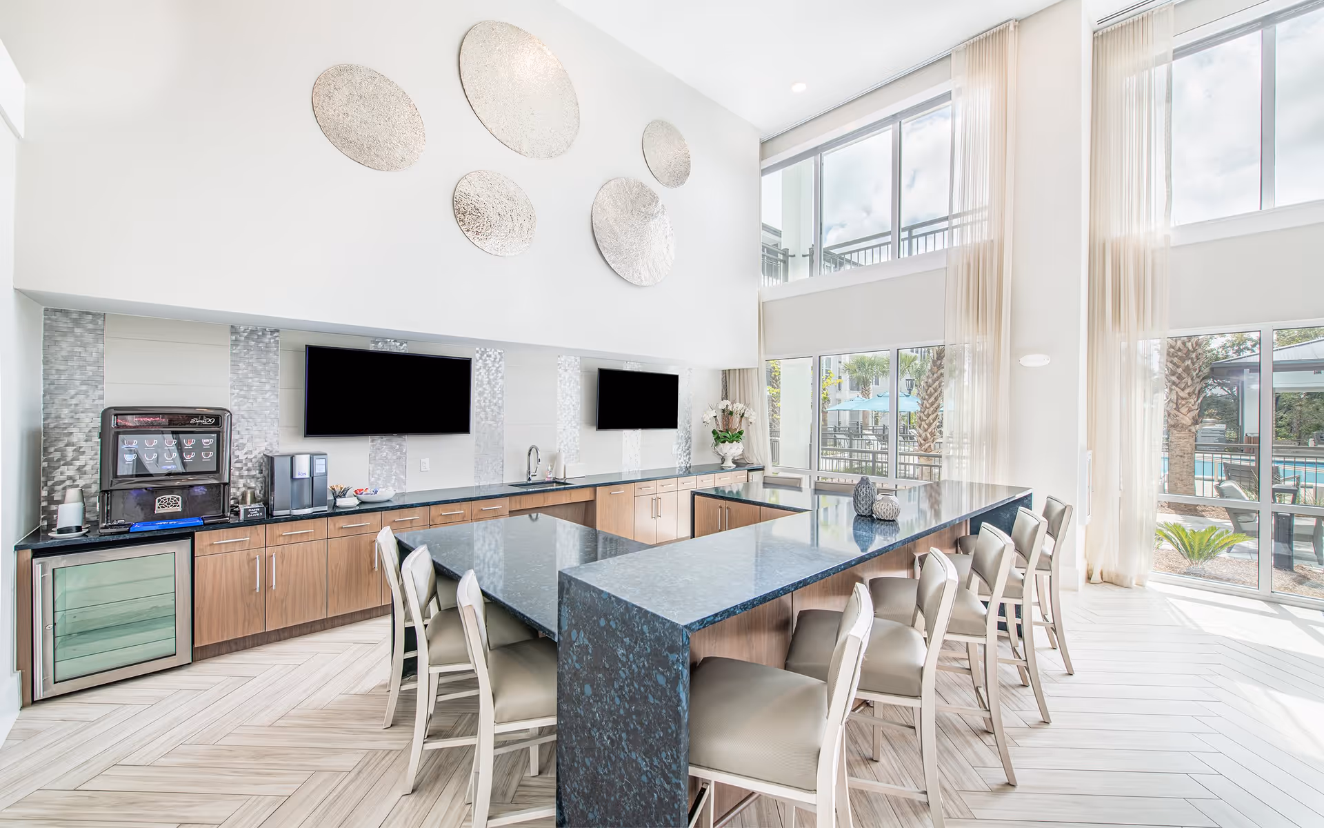 Bright modern communal kitchen and dining area with a large dark island surrounded by bar stools, wall-mounted TVs, coffee station, and floor-to-ceiling windows overlooking the pool.