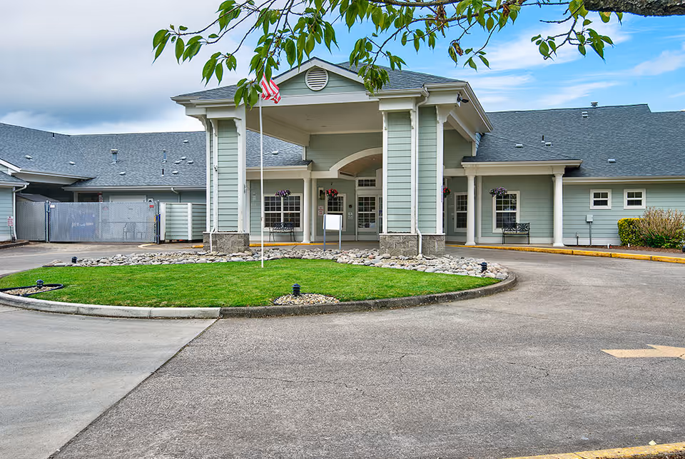 Front exterior view of a single-story assisted living facility building with a covered entrance supported by columns, an American flag on a flagpole in front, green lawn and landscaping with rocks, and a paved driveway leading to the entrance.