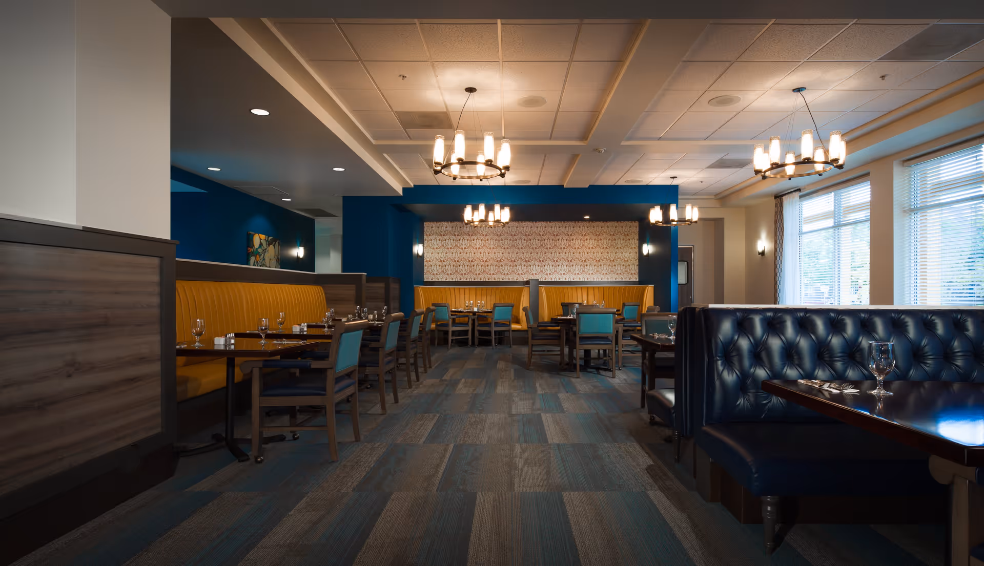 Interior view of a dining room in Russellville Park facility featuring wooden tables set with glassware and silverware, blue and yellow upholstered seating booths, blue walls, large windows with blinds, and modern chandeliers hanging from the ceiling.