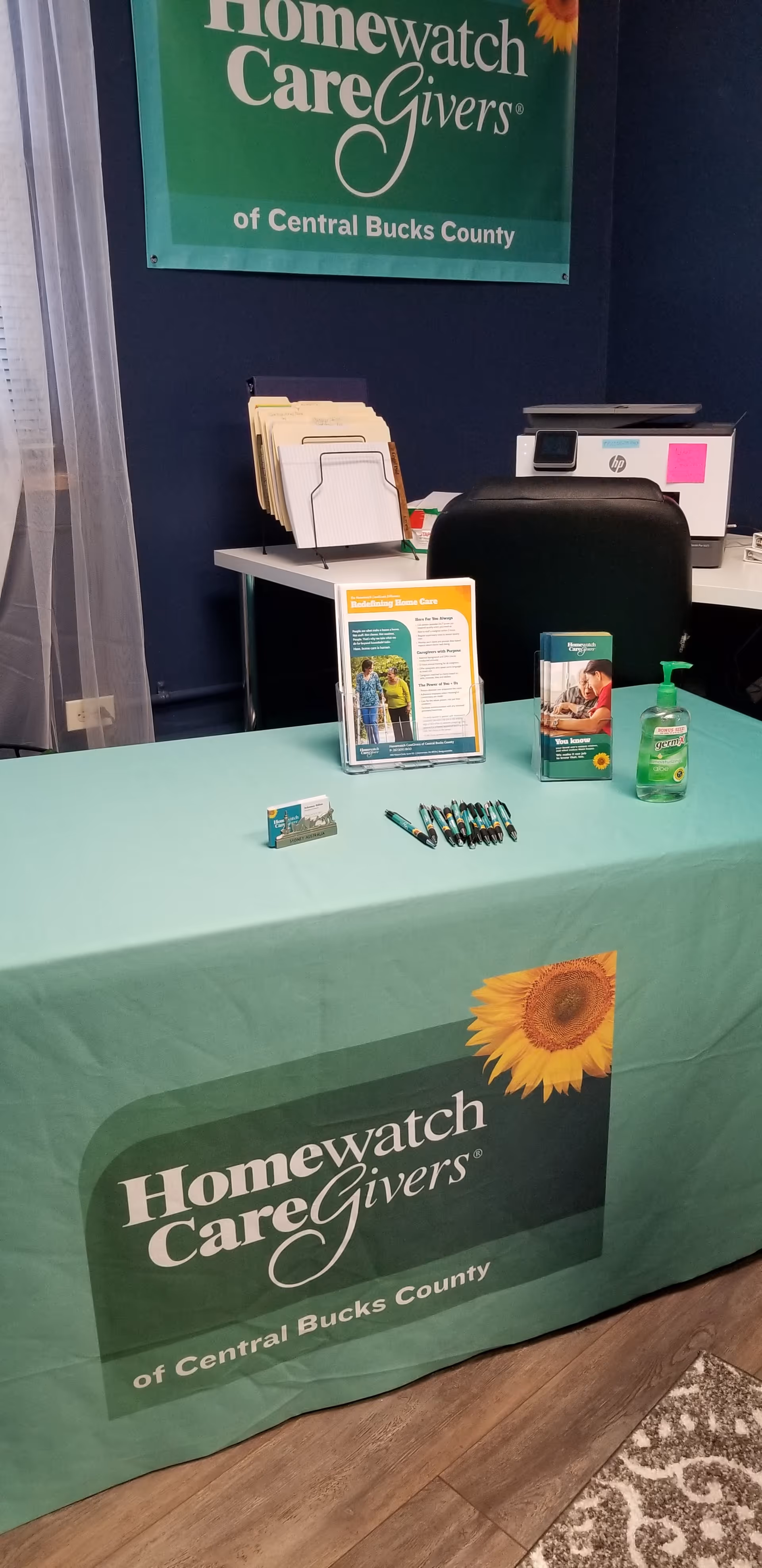 Promotional table with Homewatch CareGivers banner, brochures, and sanitizer in an indoor office.