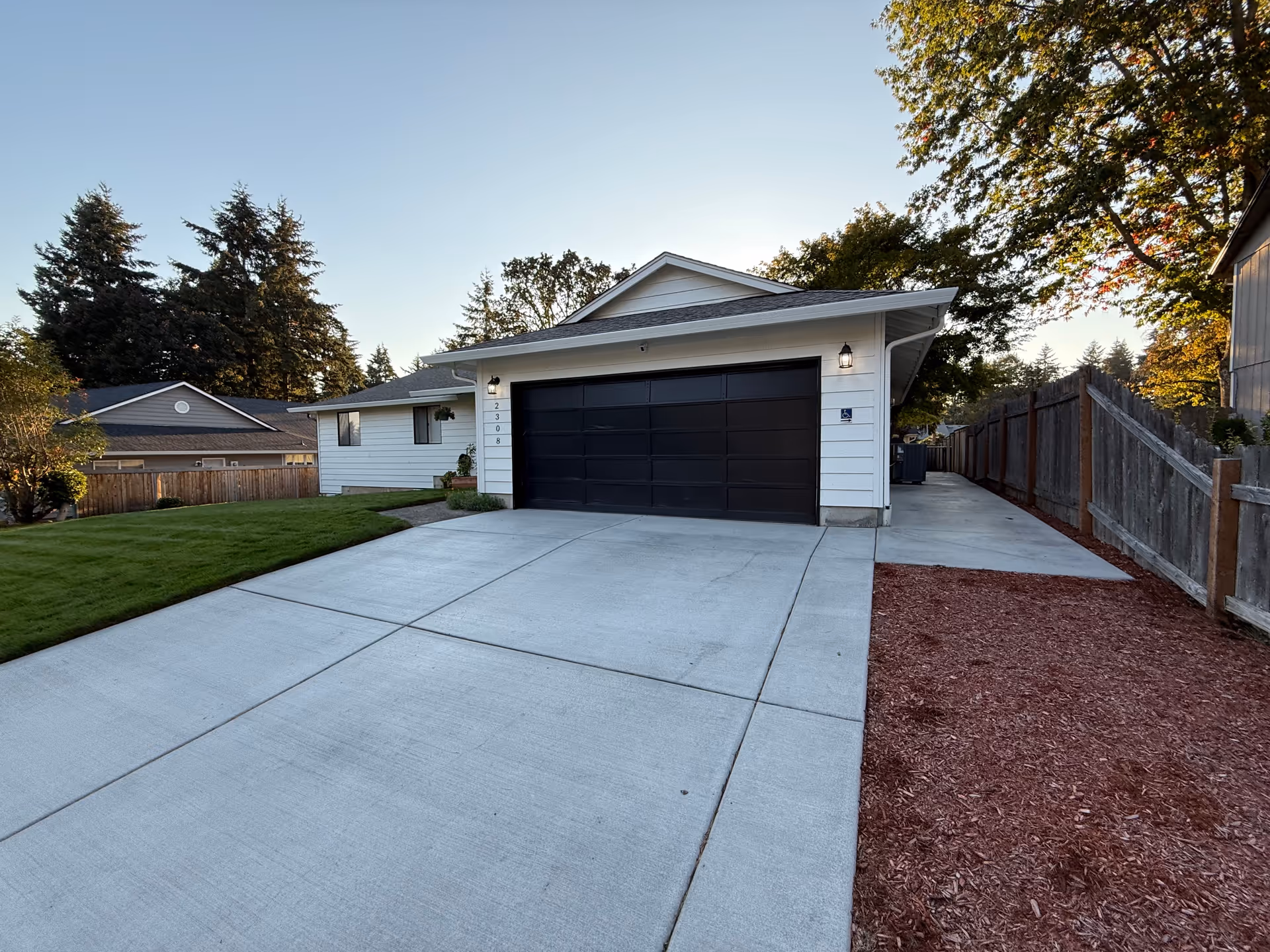 Residential single-story home with a large driveway, double garage doors, and a wooden fence.