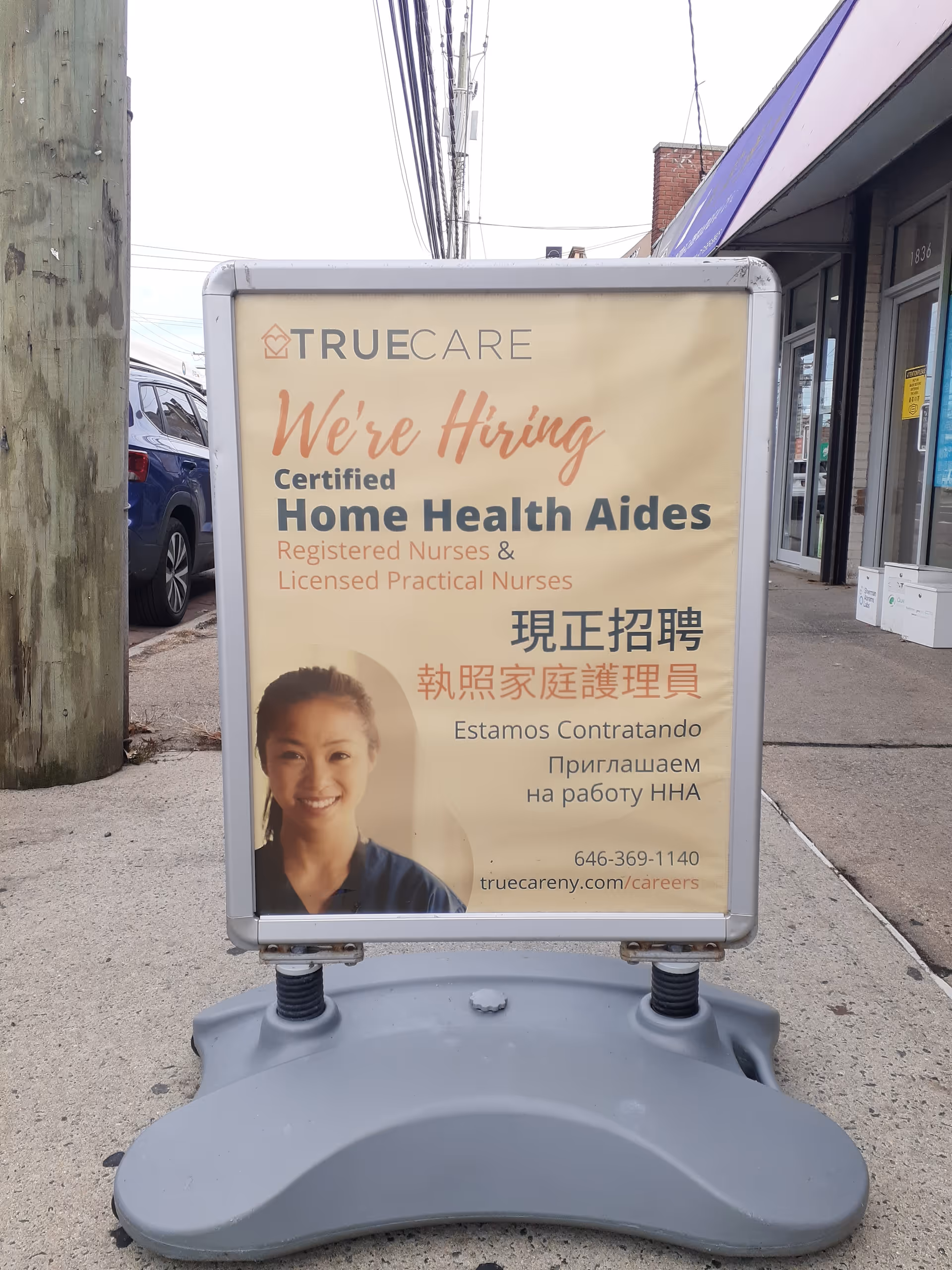 Sidewalk advertising sign for TrueCare hiring home health aides outside storefront.
