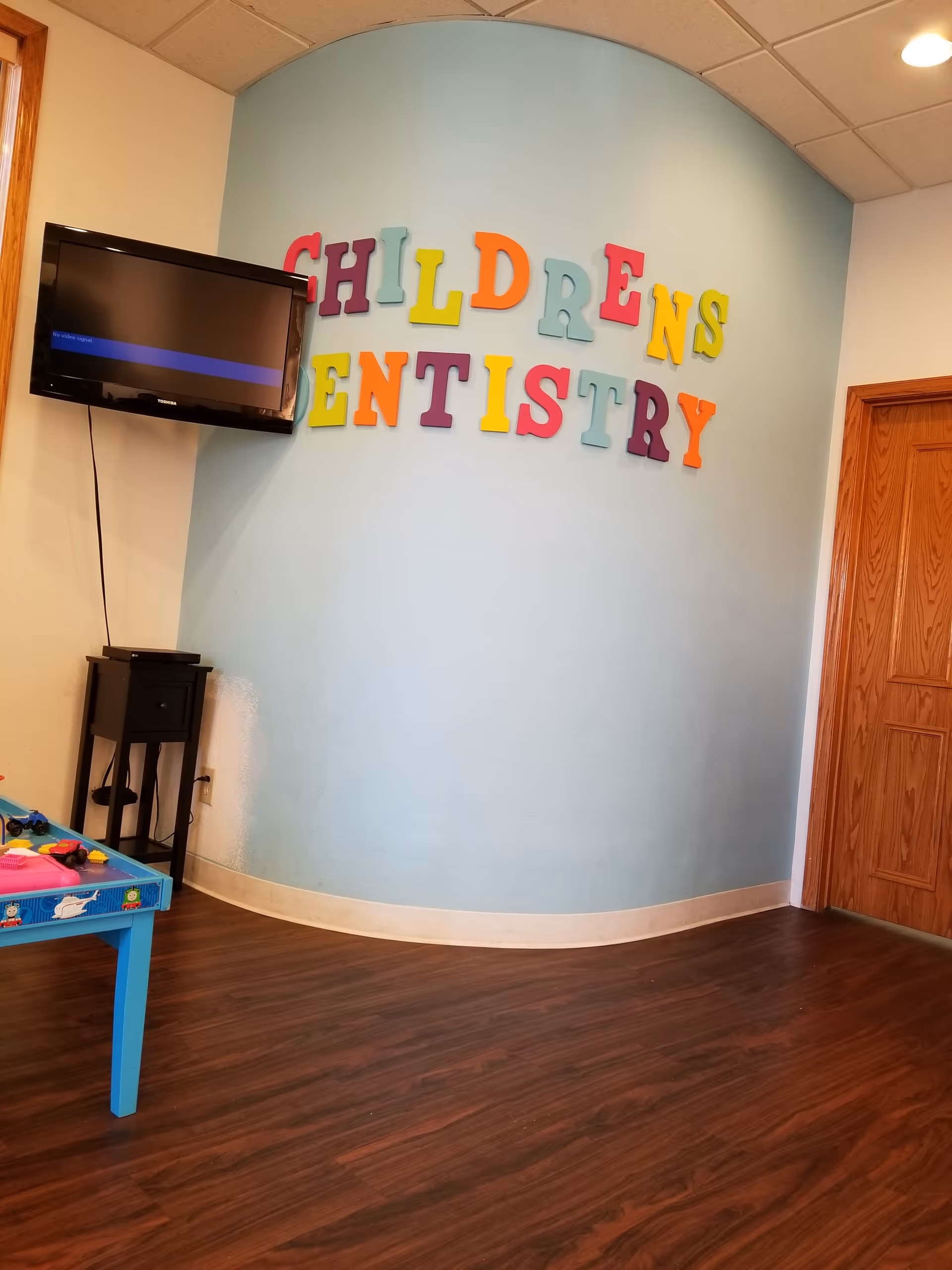 Bright pediatric play area with colorful letters spelling Children's Dentistry on a blue curved wall.