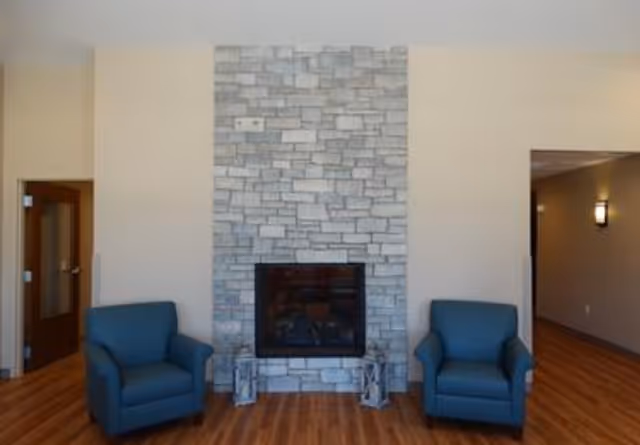A cozy interior room featuring a stone fireplace centered on a beige wall, flanked by two blue armchairs on either side. The floor is wooden, and there are doorways visible on the left and right sides of the room.