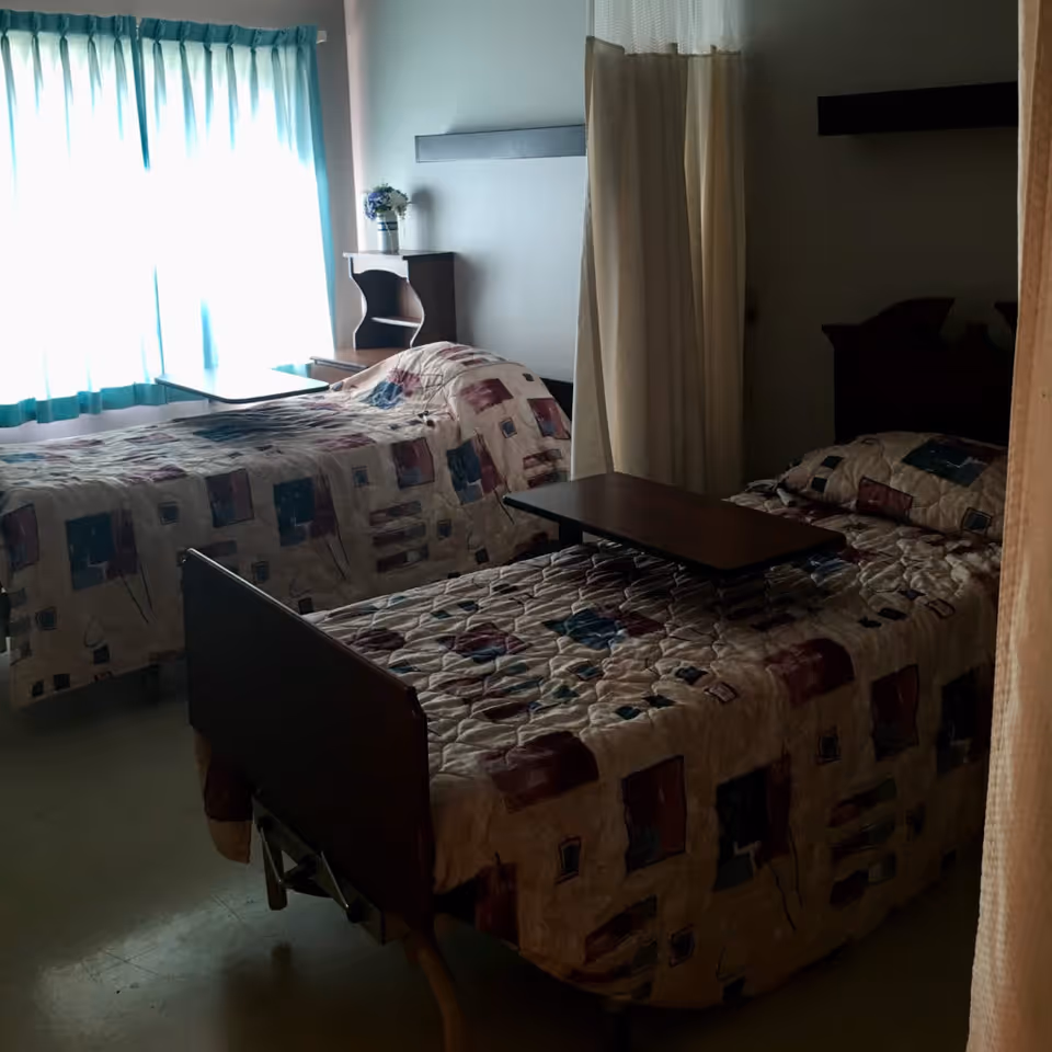A room with two hospital-style beds covered with patterned quilts, each with an overbed table. A window with light blue curtains is on the left side, and a small wooden nightstand with a vase of flowers is between the beds. A privacy curtain is partially drawn between the beds.