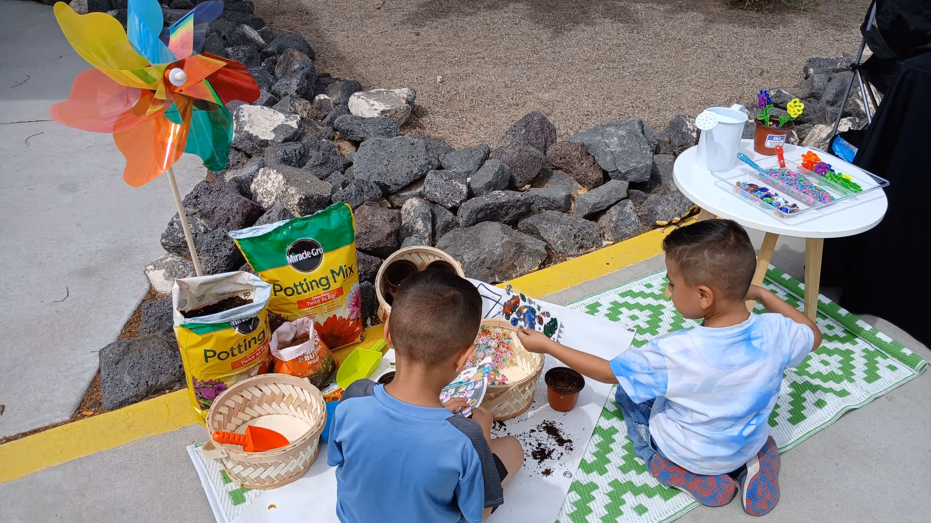 Two kids plant in pots beside a table of crafts outdoors.