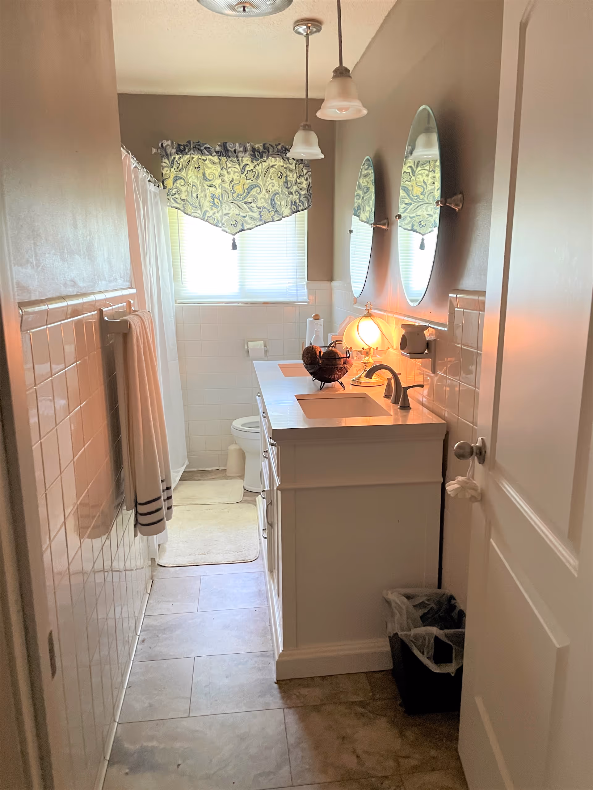 Small bathroom with white vanity, mirrors, sink, toilet, shower curtain, and window valance.