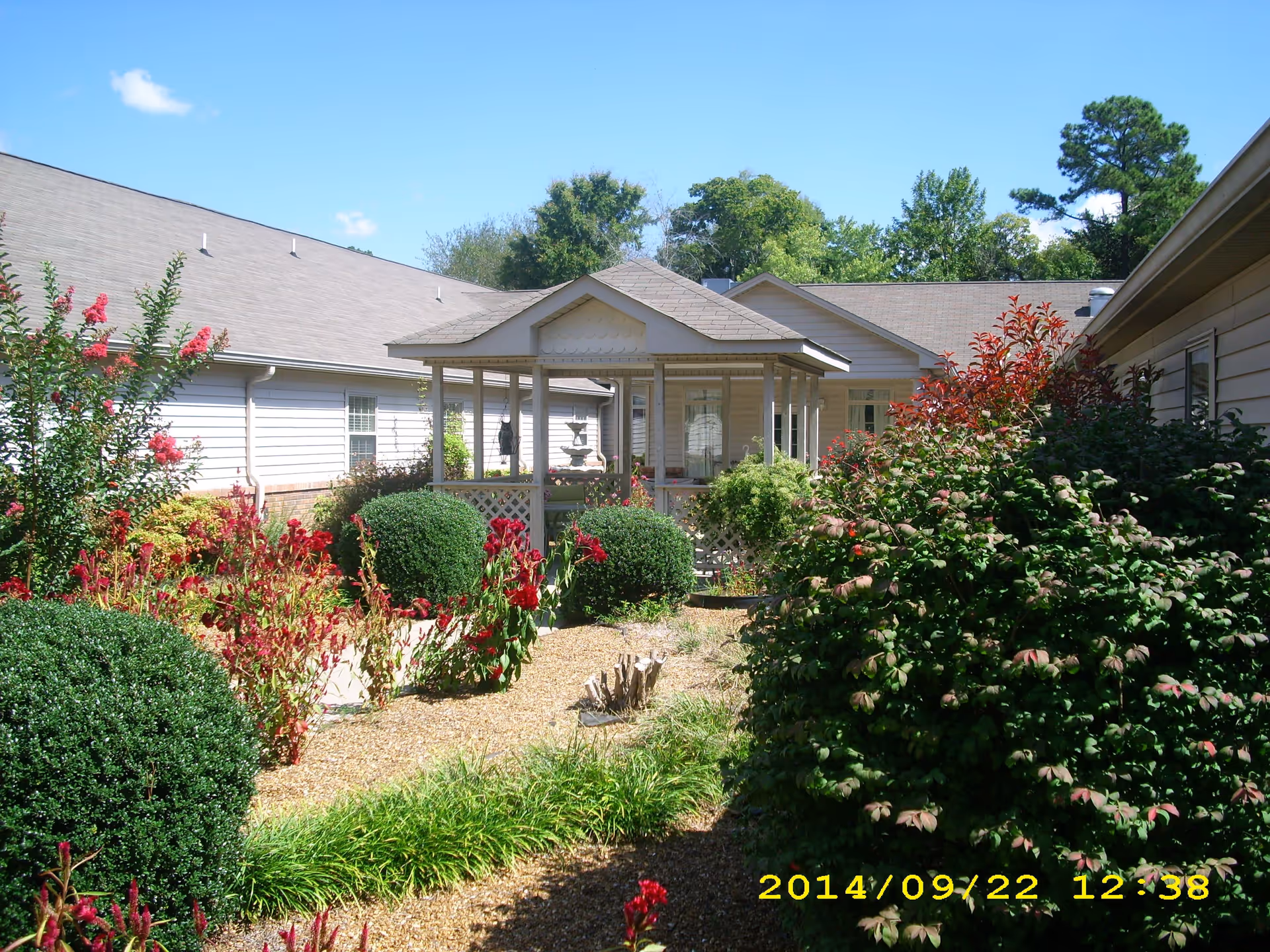 A landscaped outdoor garden area at Magnolia Place Assisted Living featuring a variety of green shrubs and red flowering plants. In the center, there is a small gazebo structure with a roof, surrounded by the garden. The building's exterior walls and windows are visible in the background under a clear blue sky.
