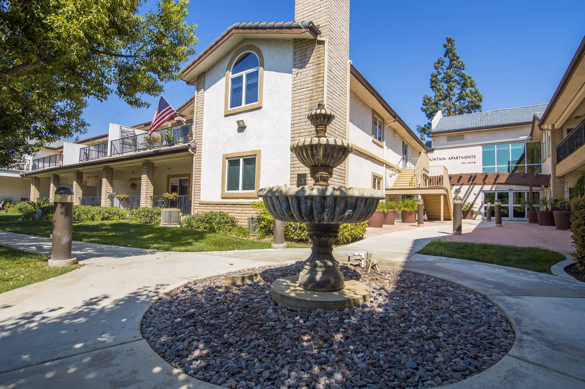 A tiered stone fountain in a courtyard in front of a two-story senior living building with balconies and an American flag.