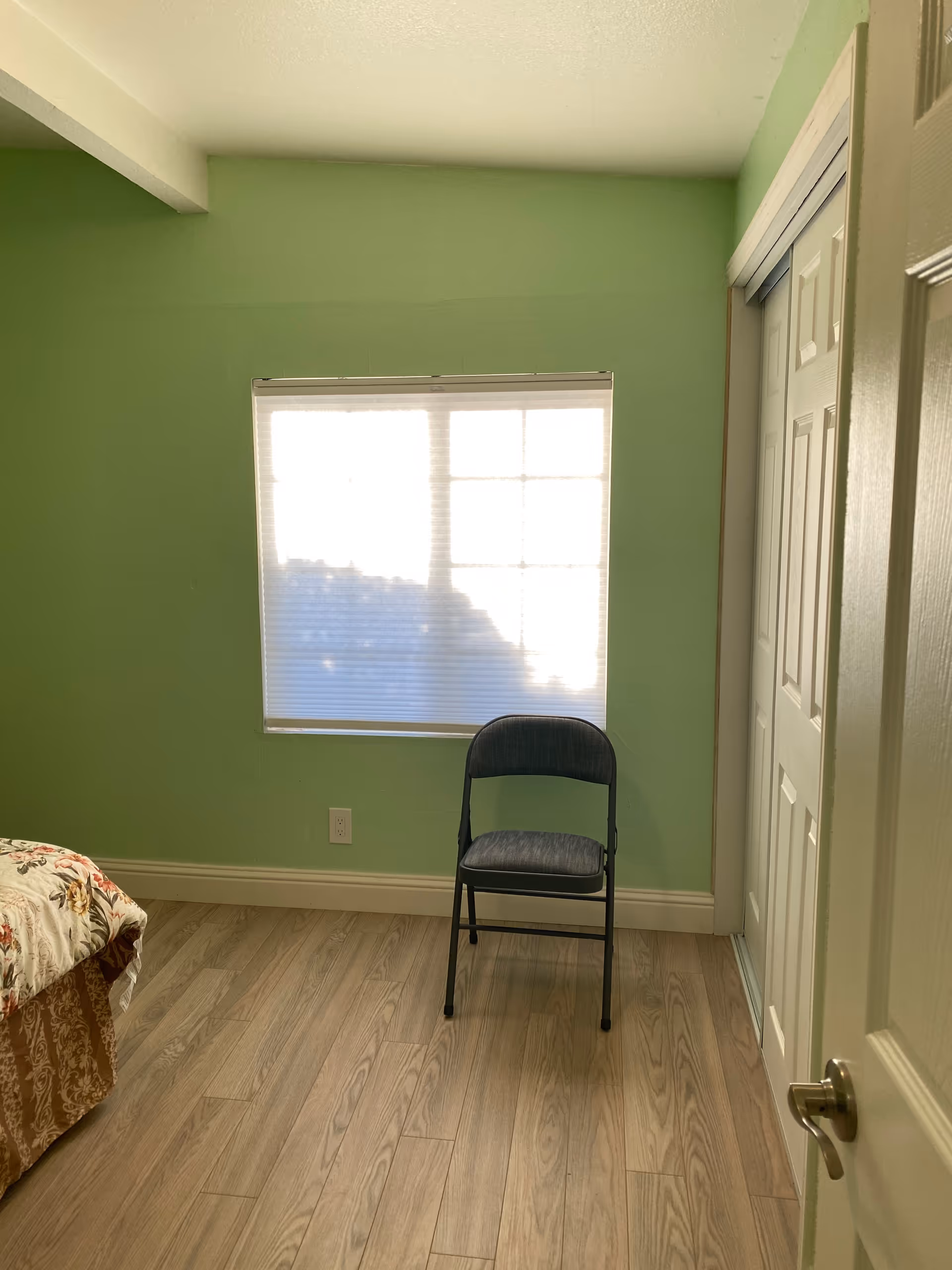 A small bedroom with light green walls and wood flooring. There is a window with blinds partially closed, a simple black folding chair placed in front of the window, and a bed with a floral patterned bedspread partially visible on the left side. A white closet with sliding doors is on the right side of the room.
