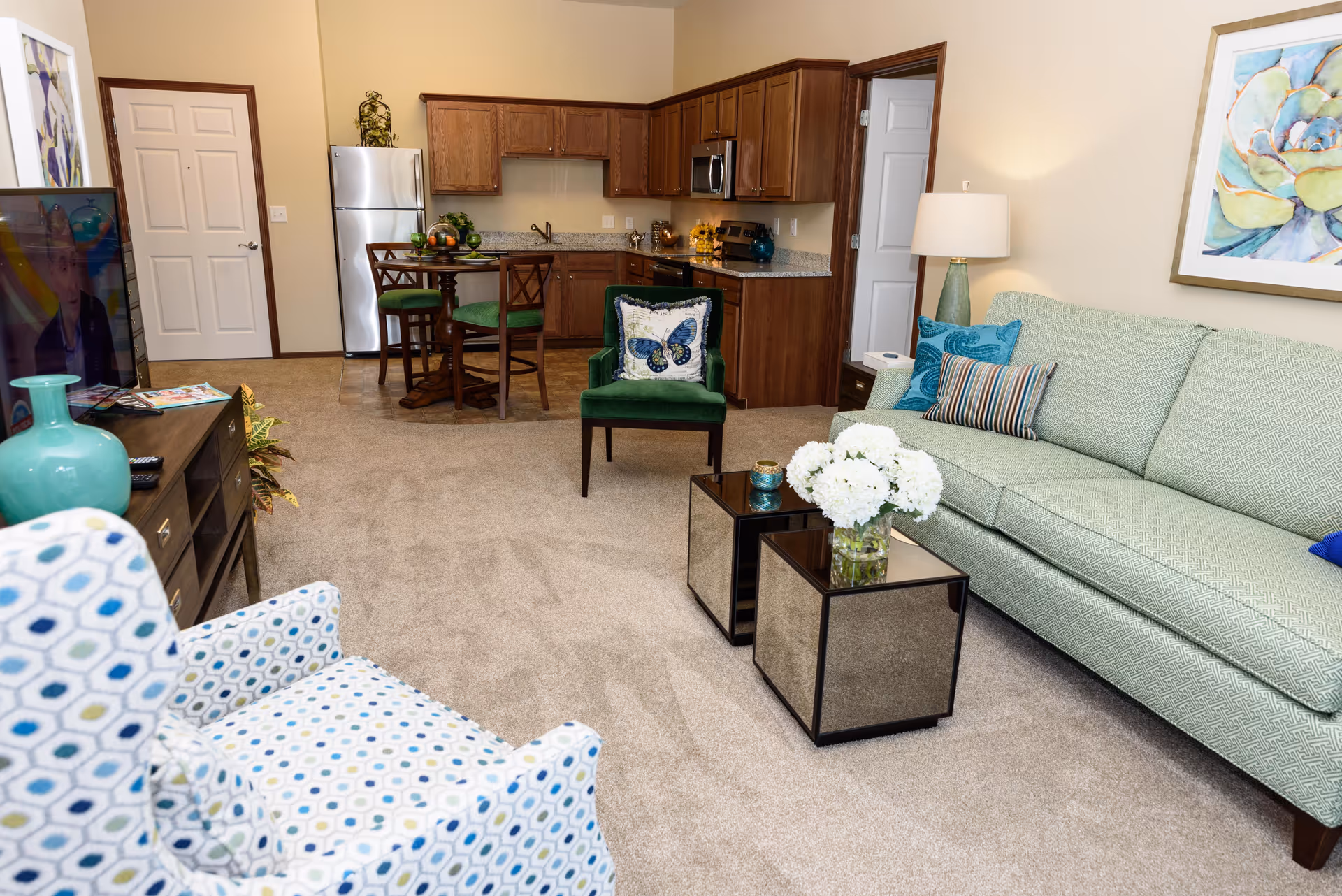 A cozy living room and kitchen area in a senior living facility. The living room features a light green sofa with colorful pillows, a patterned armchair, two mirrored coffee tables with a vase of white flowers, and a floor lamp. The kitchen area has wooden cabinets, a stainless steel refrigerator, microwave, and stove, with a small round dining table and four chairs. The walls are beige, and there is a colorful painting above the sofa.