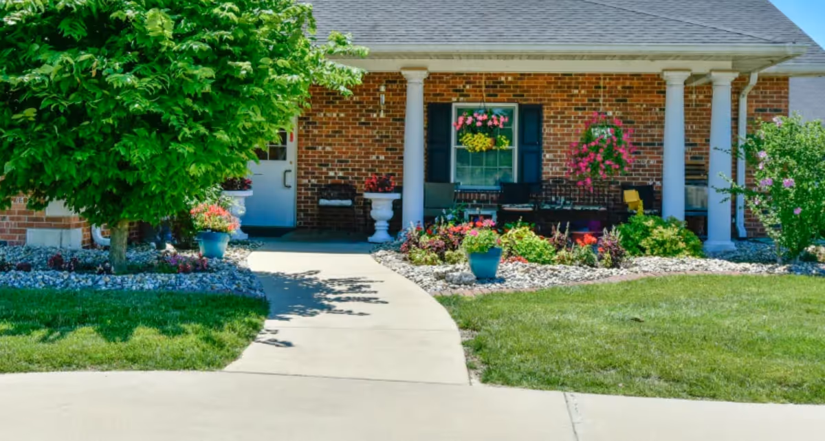 Brick senior living building with a covered front porch, walkway, and flower-filled landscaping.