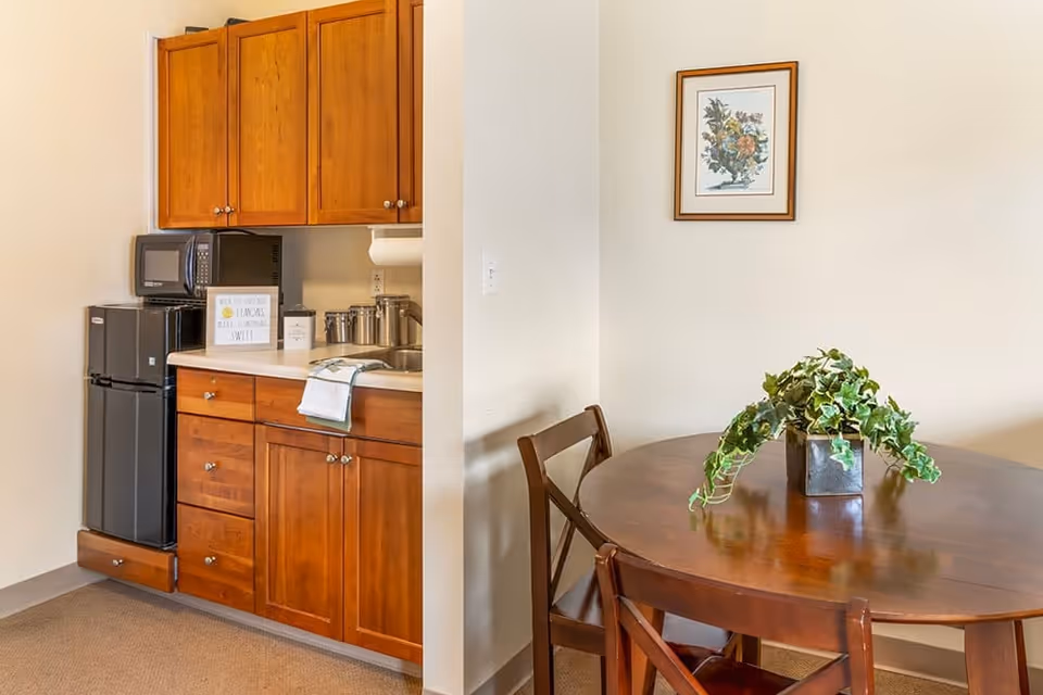 Small kitchenette with wooden cabinets, a mini-fridge and microwave next to a round wooden dining table with a potted plant.