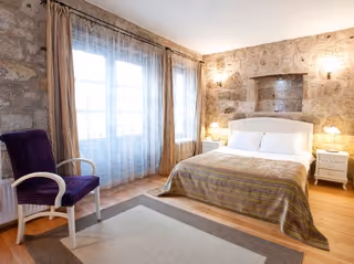 Stone-walled bedroom with a large bed, white bedding, nightstands, lamps, a purple chair, and curtains.