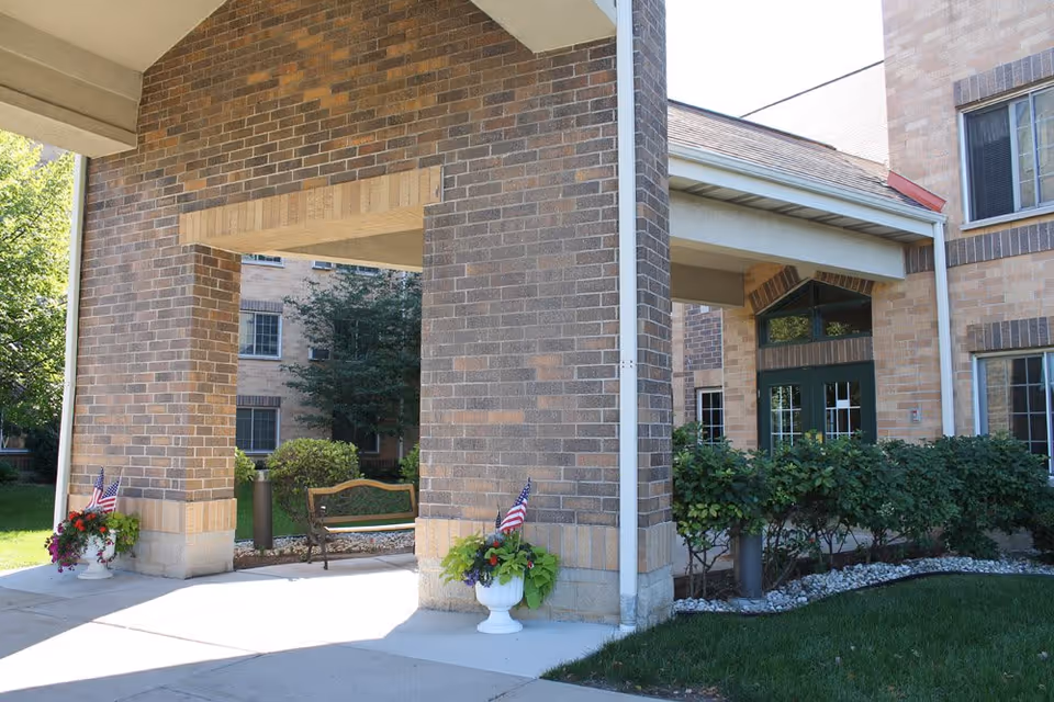 Covered brick entrance/porte-cochere of a senior living building with potted flowers, American flags, a bench, and shrubs.