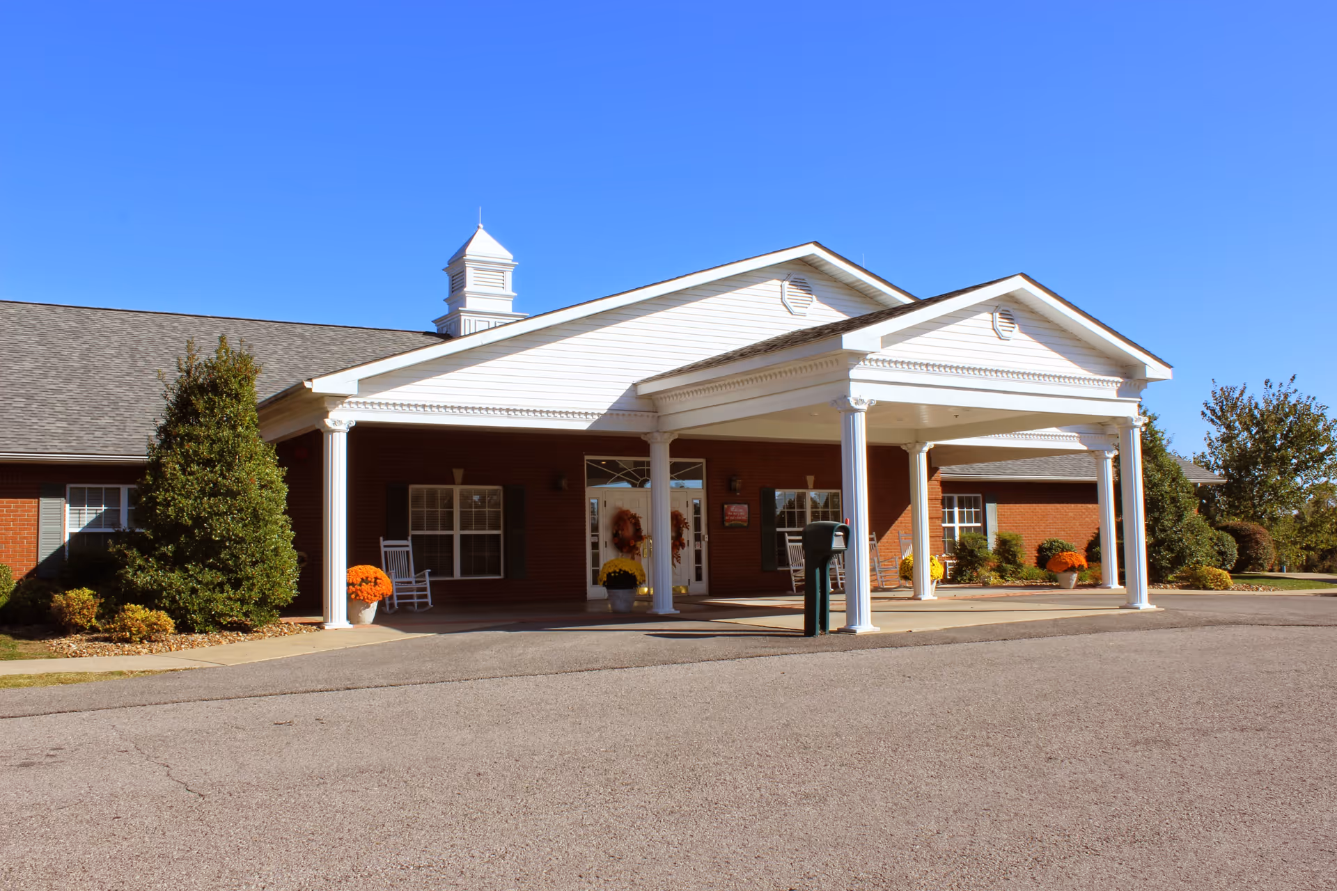 Front exterior view of Oak Ridge Senior Living Community building with a covered entrance supported by white columns, brick walls, windows with green shutters, and decorative plants and flowers near the entrance under a clear blue sky.