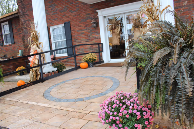 Front entrance of a brick building with a paved circular pattern, an accessibility ramp, potted chrysanthemums, pumpkins, and cornstalk decorations.