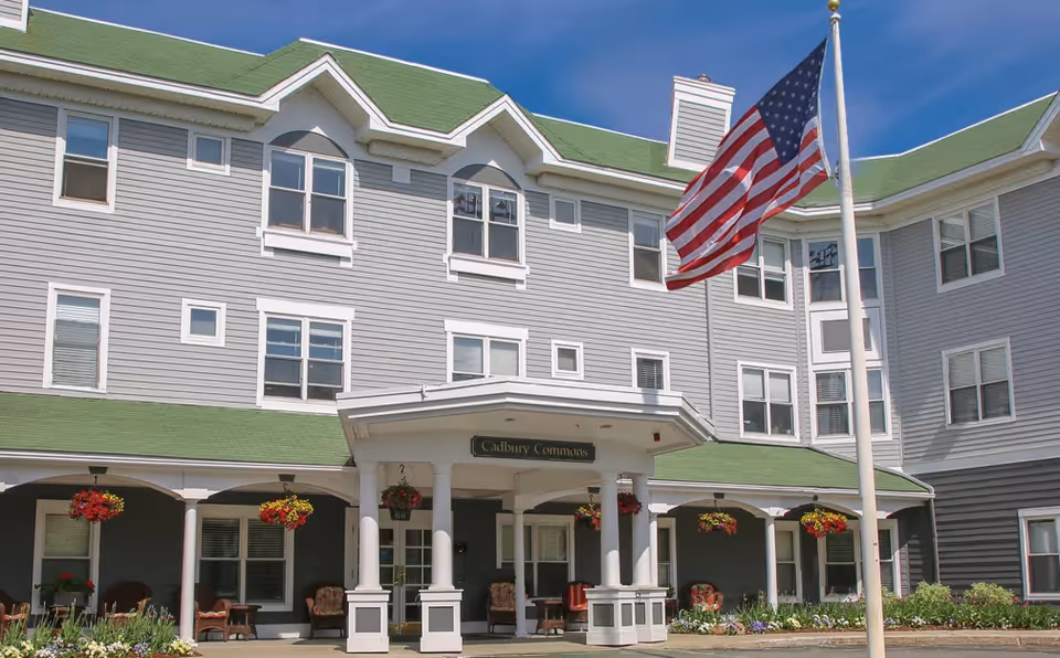 Front entrance of Cadbury Commons senior living building with a covered porte-cochere, hanging flower baskets, and an American flag flying on a flagpole.