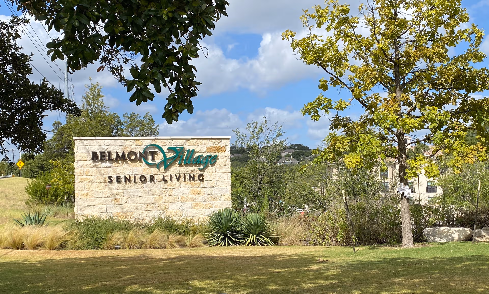 Stone Belmont Village Senior Living sign with manicured landscaping and trees in front.