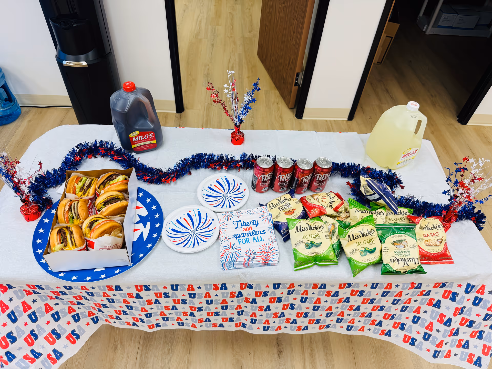 Table set indoors with hot dogs, chips, sodas, and patriotic decorations.