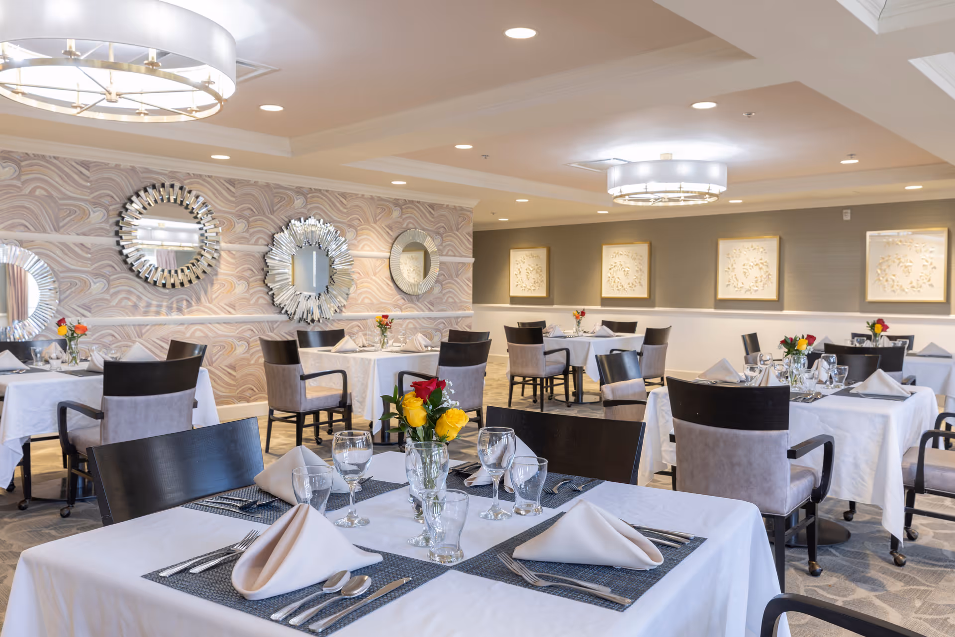 A bright and elegant dining room with multiple tables covered in white tablecloths, each set with folded napkins, glassware, and silverware. Chairs with dark wooden backs and light cushioned seats surround the tables. The walls feature decorative mirrors and framed artwork, and modern ceiling lights illuminate the space.