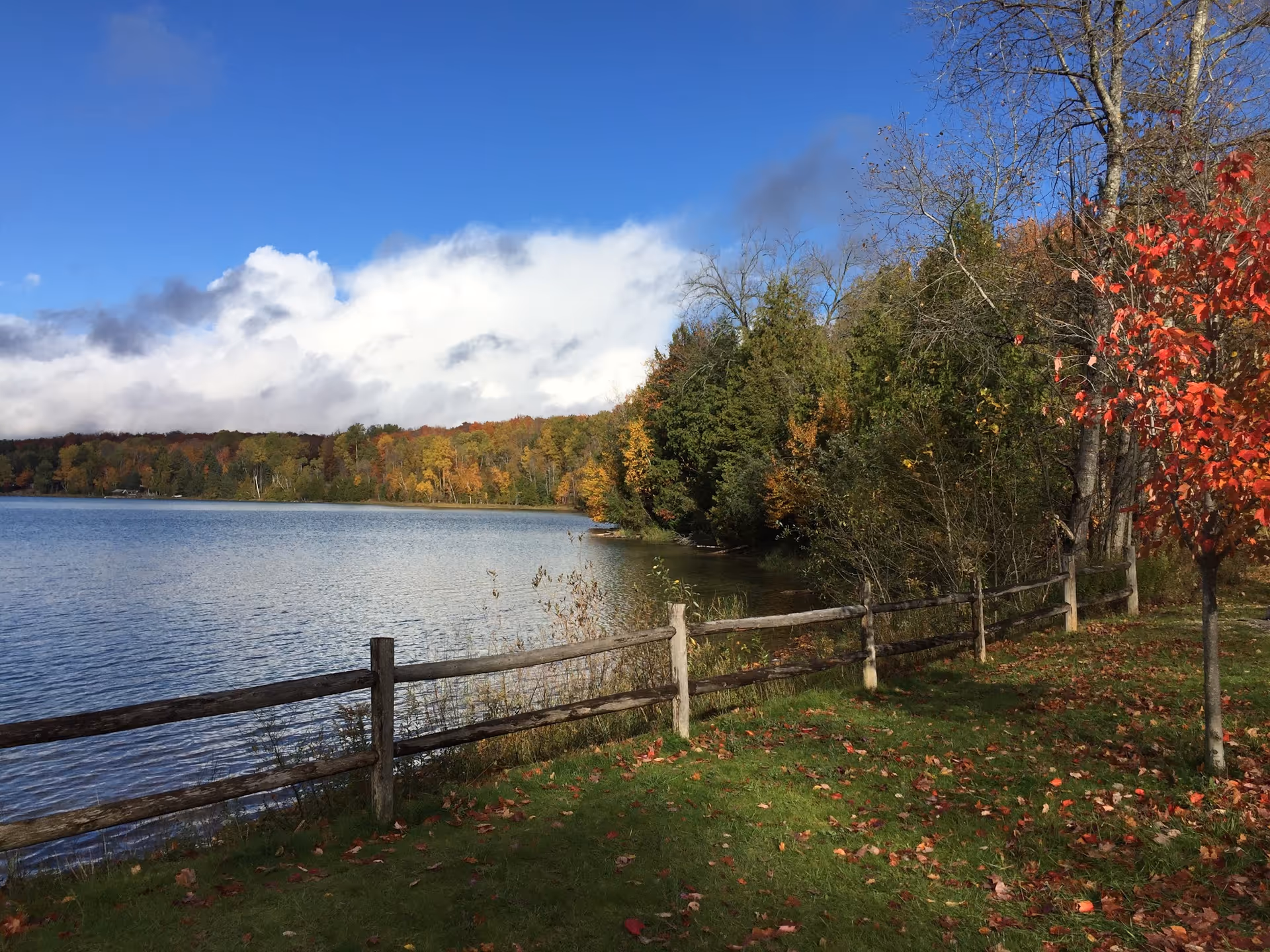 Lakeside autumn scene with a wooden fence and colorful trees.
