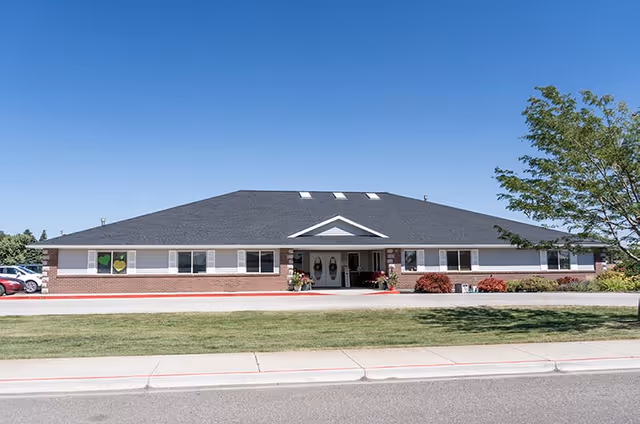 Single-story brick and siding assisted living building with a wide gabled roof, central entrance canopy, and a manicured lawn under a clear blue sky.