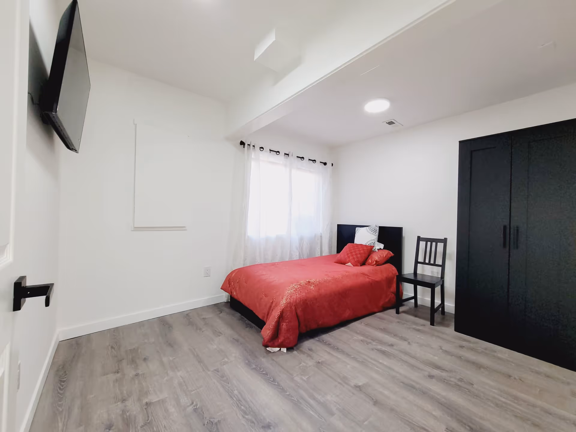 A minimalist bedroom with a red bedspread, black wardrobe, chair, window, and mounted TV.