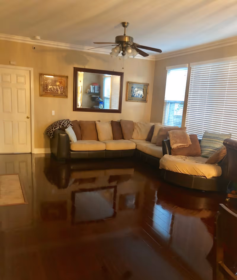 A cozy living room with a large L-shaped sectional sofa in beige and brown tones. The room has polished dark wooden floors, beige walls, two framed pictures, a large mirror on the wall, and two windows with white blinds. A ceiling fan with lights hangs from the ceiling.