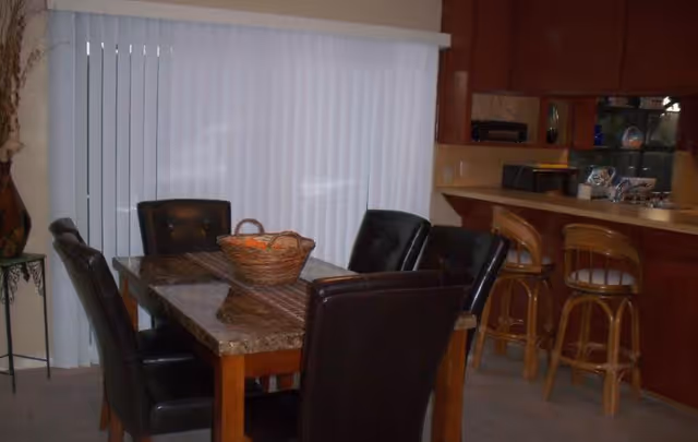 Dining area with a marble-top table surrounded by dark leather chairs, a wicker basket centerpiece, vertical blinds, and a kitchen counter with three barstools.
