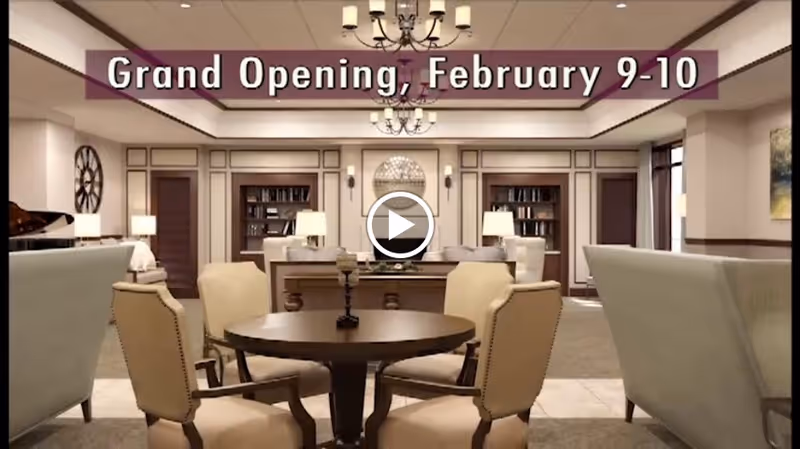 A spacious and elegantly furnished senior living common area with beige armchairs around a round wooden table in the foreground, bookshelves and lamps in the background, and a chandelier hanging from the ceiling. Text overlay reads 'Grand Opening, February 9-10'.