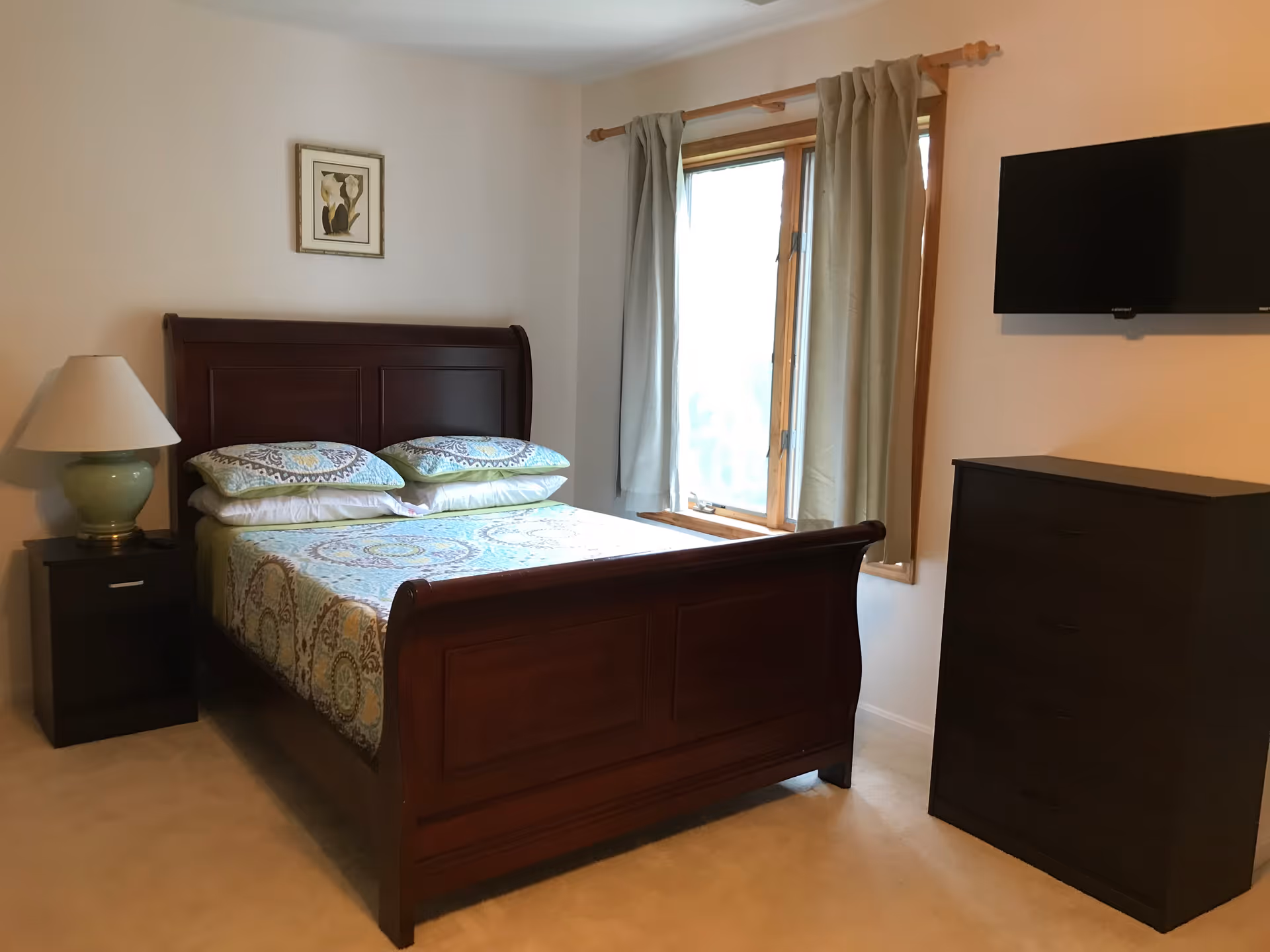 Bedroom featuring a wooden sleigh bed with patterned bedding, a nightstand and lamp, a dresser with a wall-mounted TV, and a curtained window.