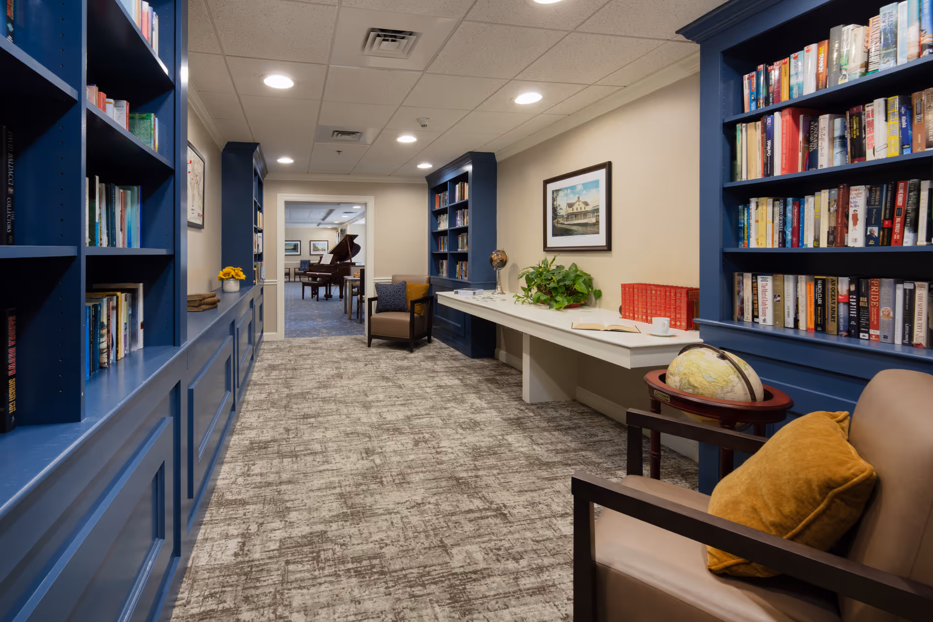 A cozy library hallway with blue bookshelves filled with books on both sides. There are two armchairs with mustard-colored pillows, a small globe on a stand, a white desk with a plant, books, and a cup. The hallway leads to a room with a grand piano and more seating. The walls are beige, and the carpet is patterned in neutral tones.