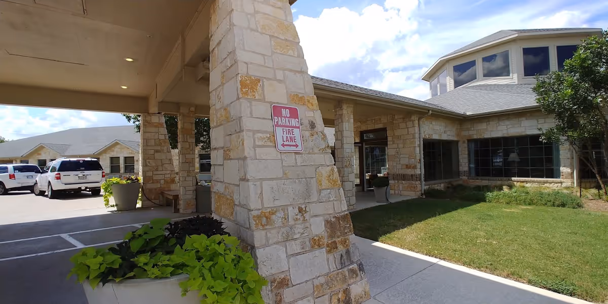 Stone‑façade nursing facility entrance with a covered porte‑cochere, parked cars, planters, and a lawn.