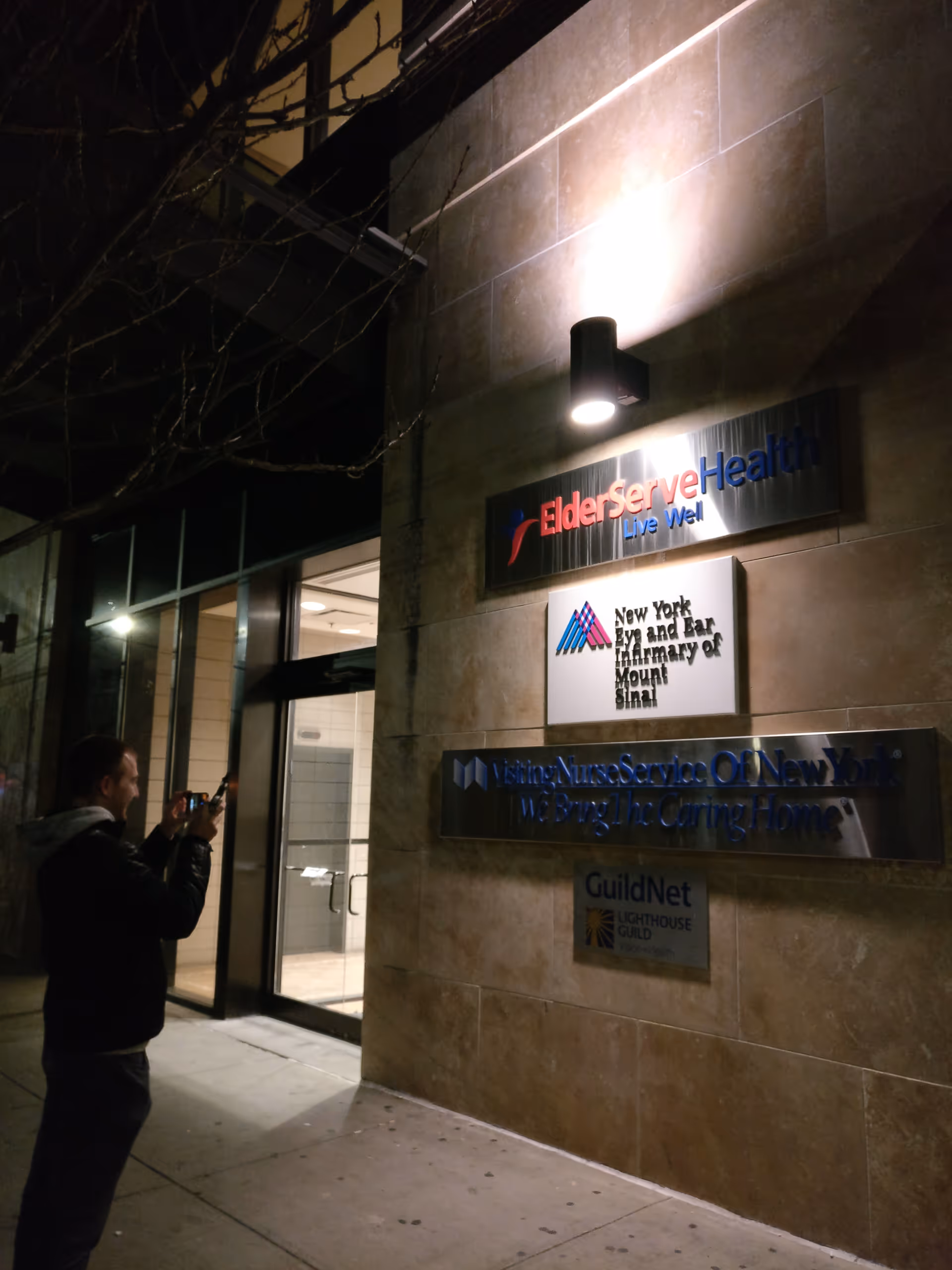 Night exterior of a building with lit signage and a person taking photos at the entrance.