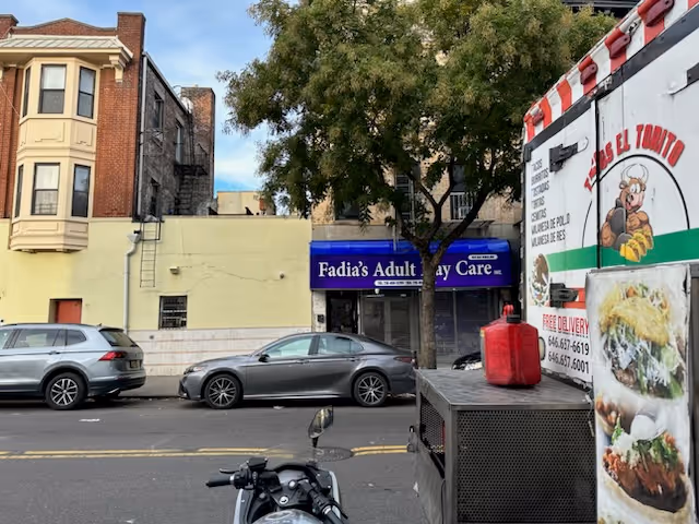 Street view with parked cars, a blue Fadia's Adult Day Care sign, and a food truck.