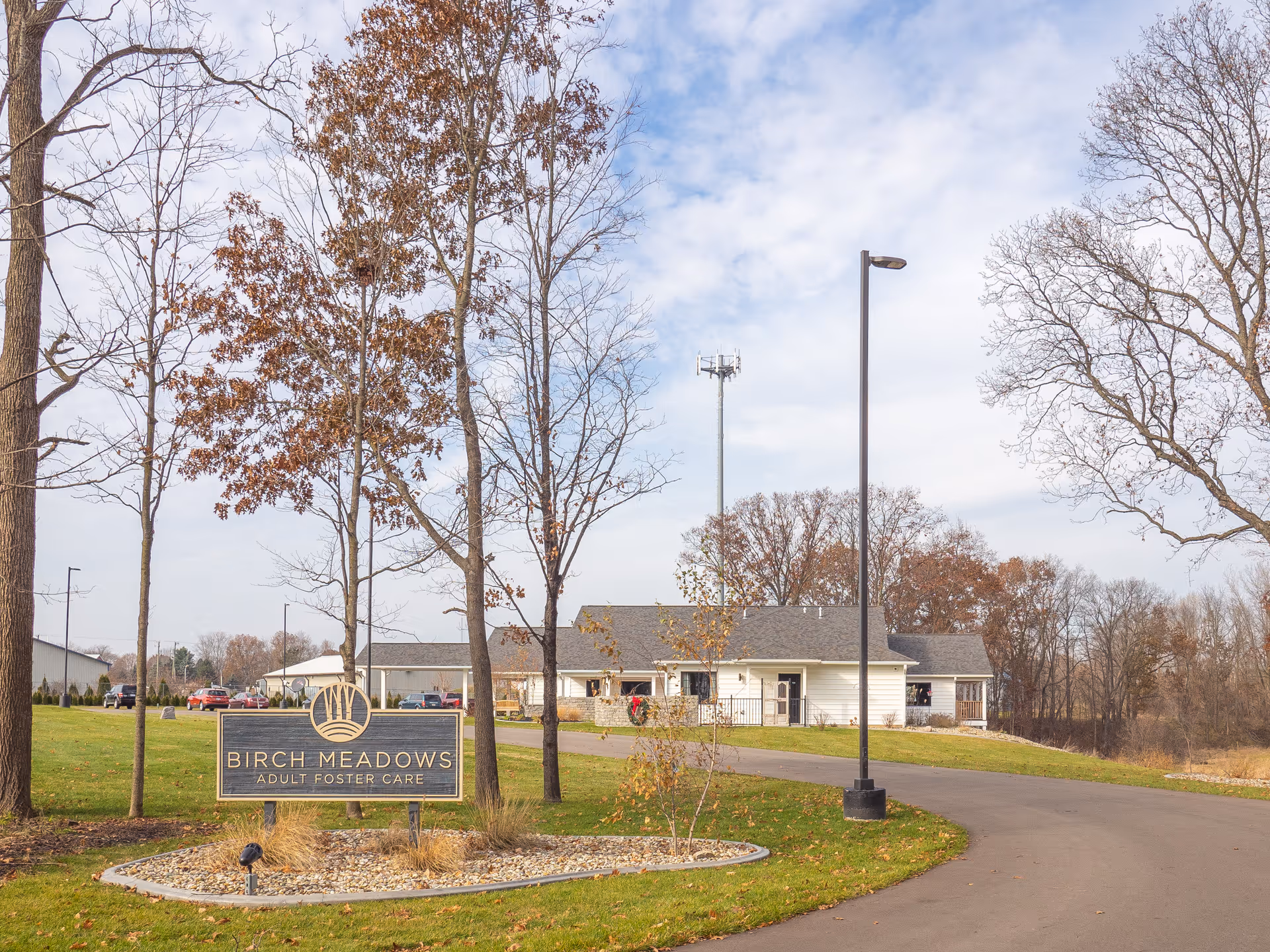 Exterior view of Birch Meadows Adult Foster Care facility with a sign in the foreground surrounded by trees with autumn leaves and a paved driveway leading to the building under a partly cloudy sky.