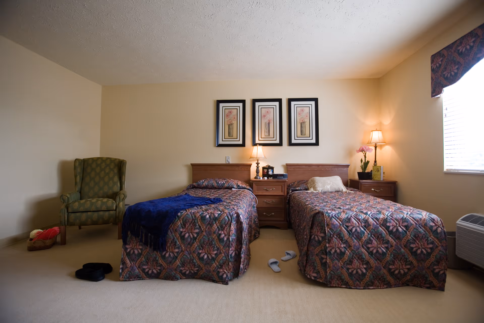 Two twin beds with matching quilts, nightstands, lamps, and an armchair in a plain bedroom.
