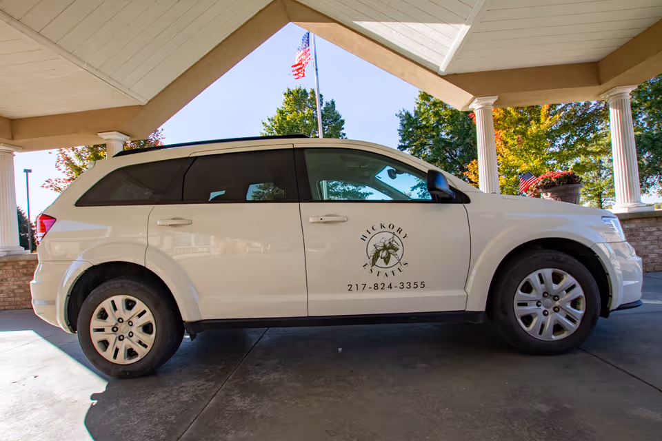 White SUV bearing a 'Hickory Estates' logo parked under the facility's covered entrance with an American flag visible behind it.