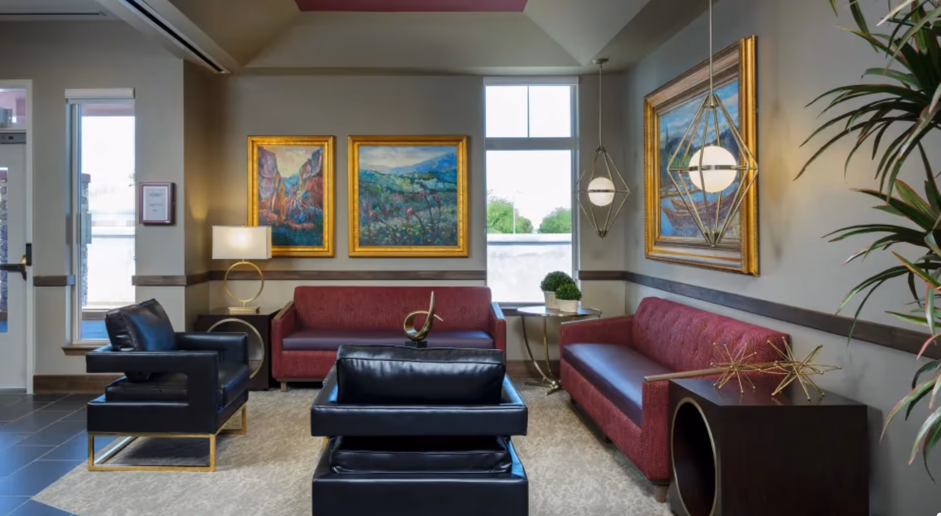 A modern living room area with two red sofas and two black leather armchairs arranged around a beige carpet. The walls are decorated with three large framed paintings and two geometric pendant lights hang from the ceiling. There is a small side table with decorative plants and a dark wooden side table with gold star sculptures. A large window lets in natural light.